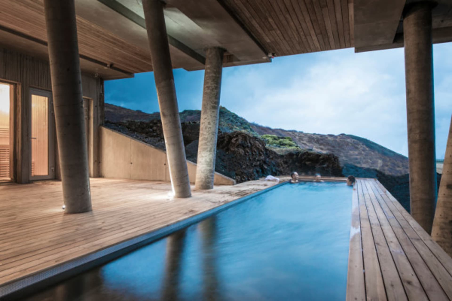 Swimming pool deck with wooden floor and concrete pillars surrounded by rocky landscape.