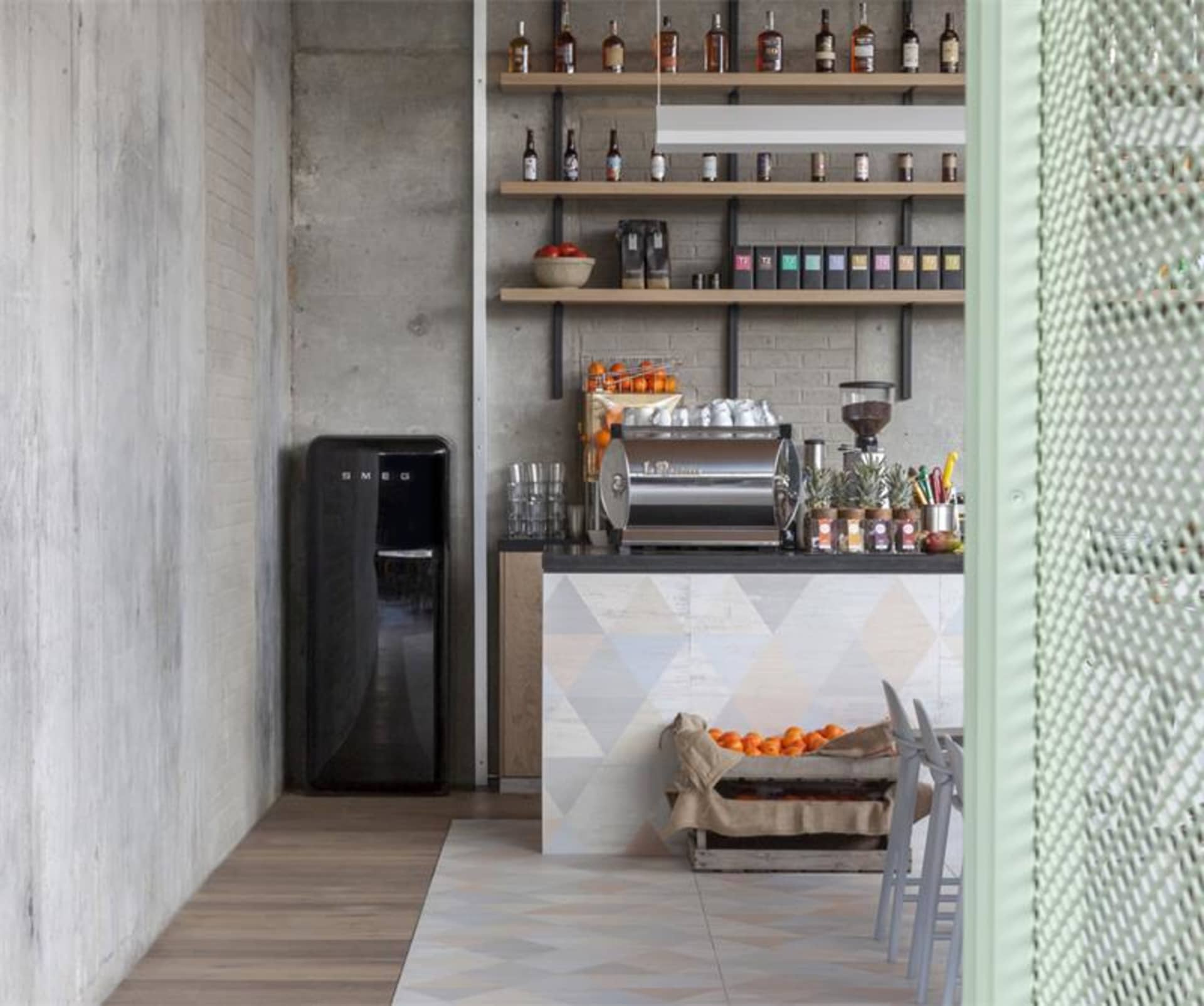 A modern industrial coffee shop with concrete shelving, featuring coffee-making equipment, oranges in a crate, and a SMEG ref