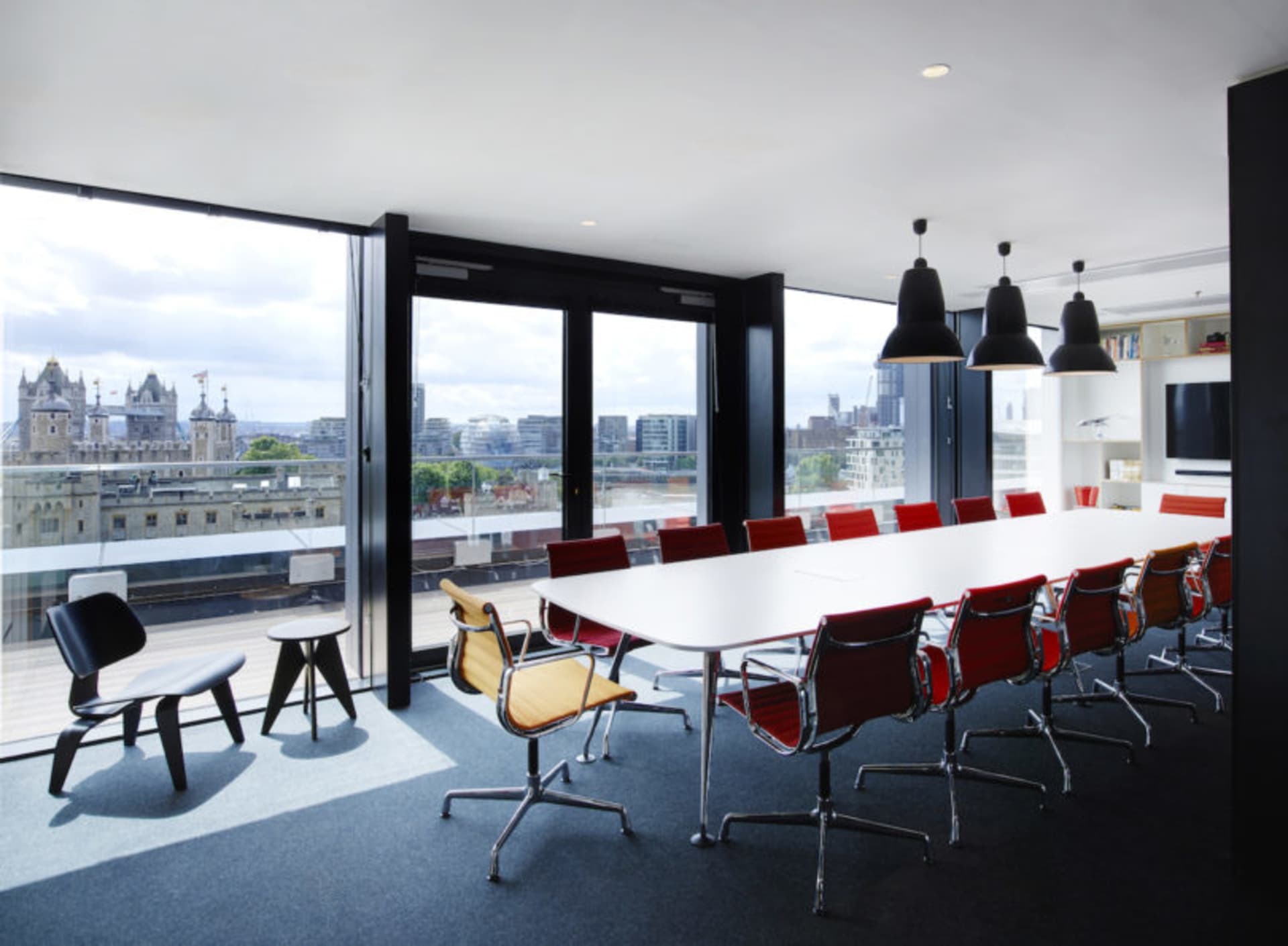 A modern conference room featuring a long white table surrounded by red chairs, black pendant lights, and a view of a citysca