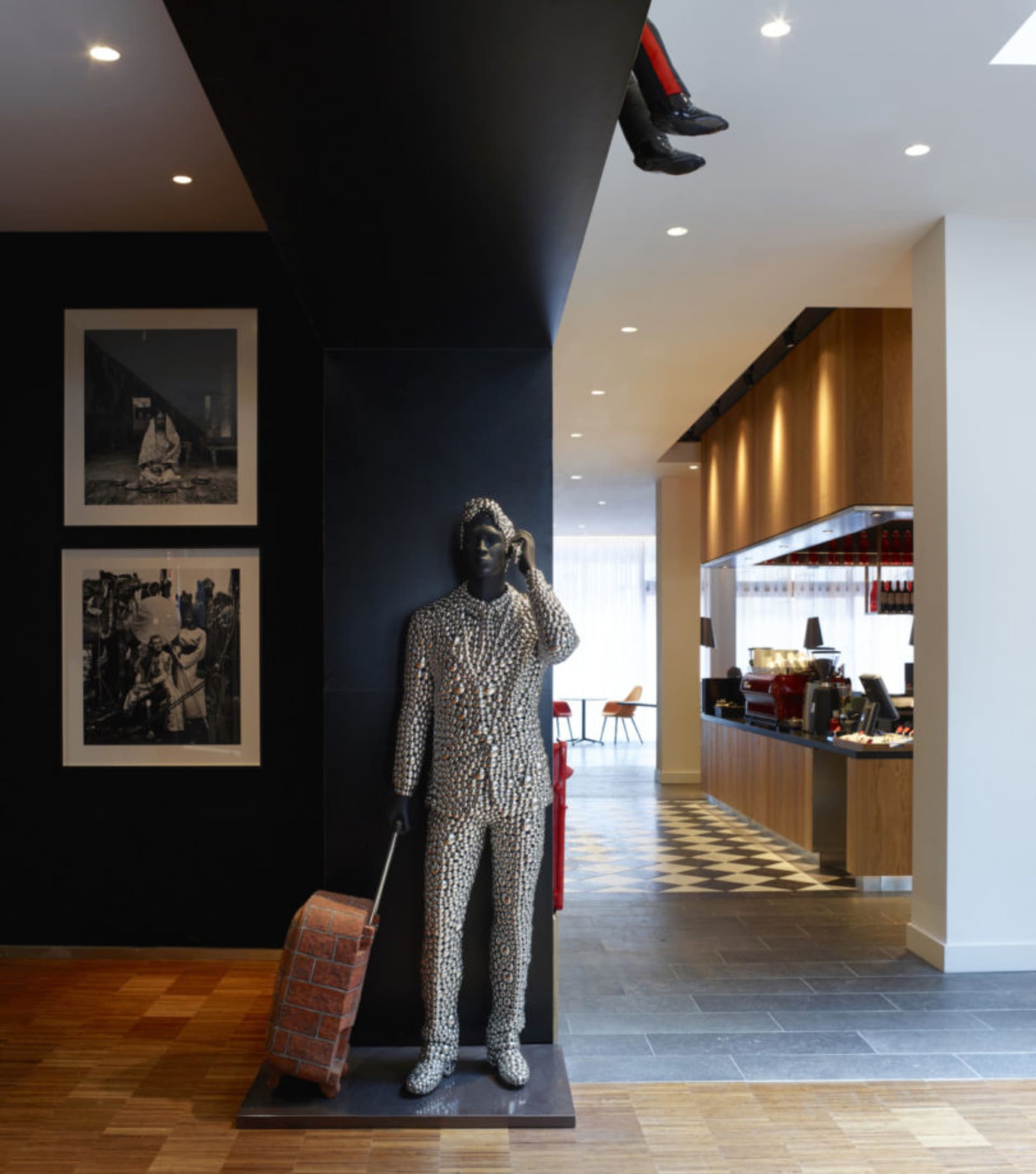 A modern statue of a person dressed in a patterned suit and holding a suitcase stands in a lobby with black and white checker