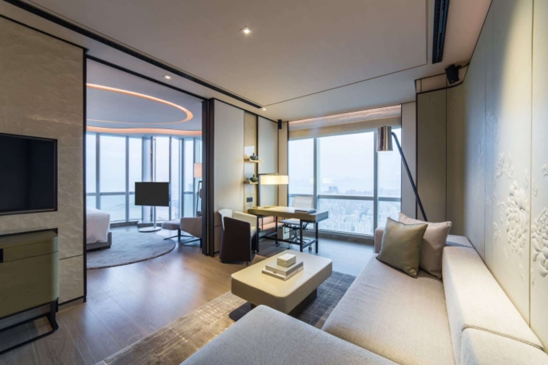 A modern minimalist living room with white decor, featuring a large sofa, coffee table, and desk area with a cityscape view.