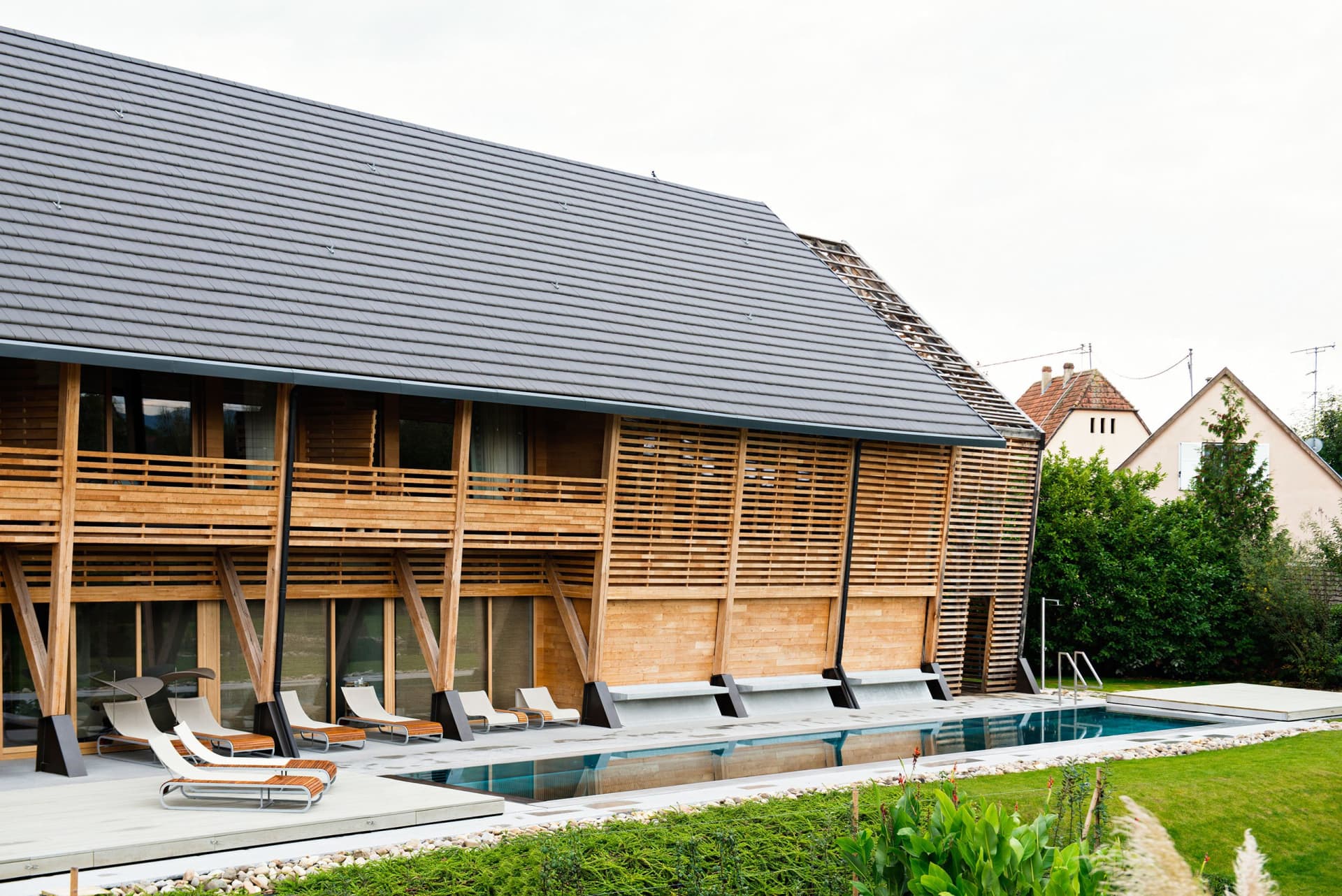 A modern residential exterior with a wooden facade and a pitched roof with a pool in the foreground.