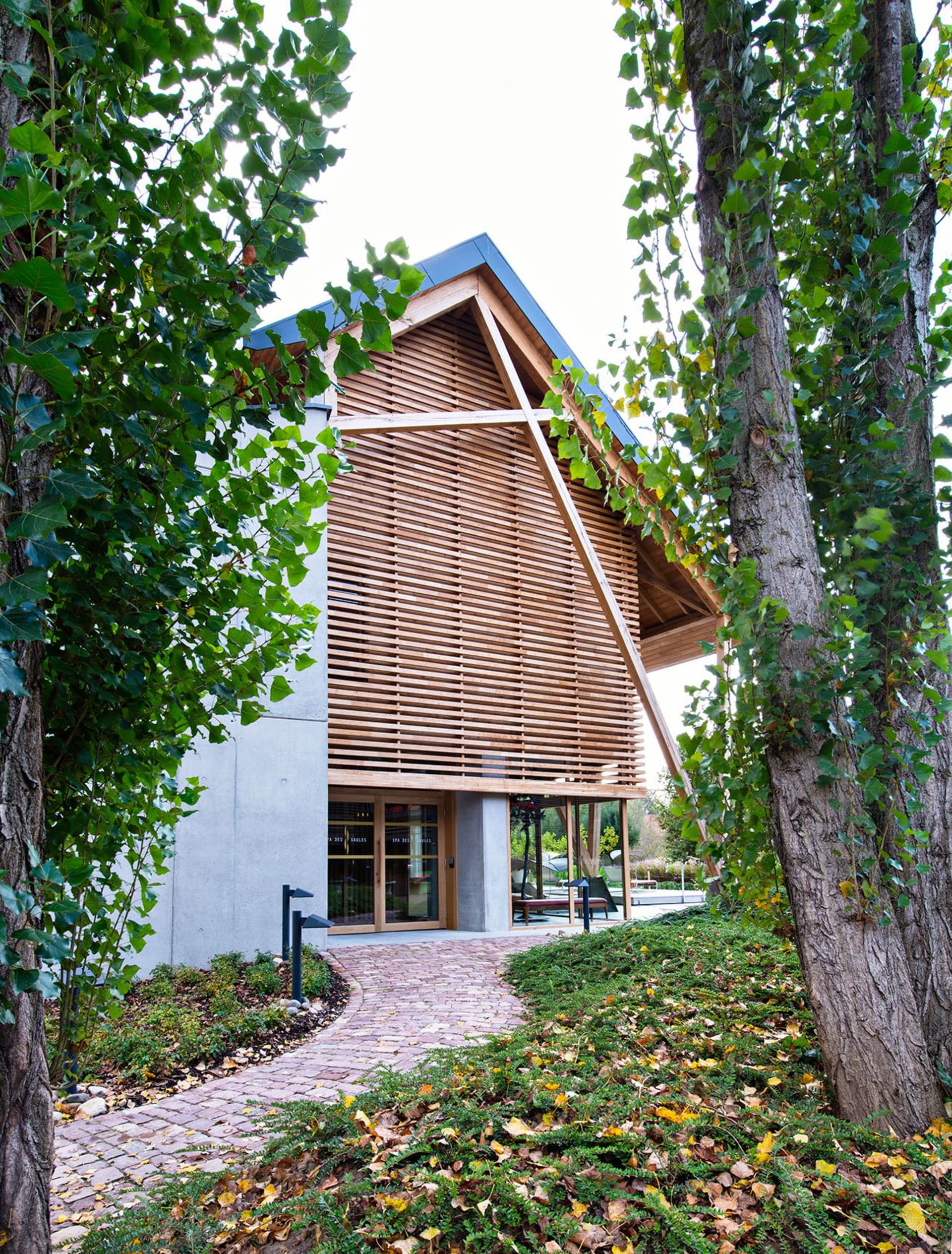 A modern building exterior made of wood and concrete with a sloping roof and large windows surrounded by greenery.