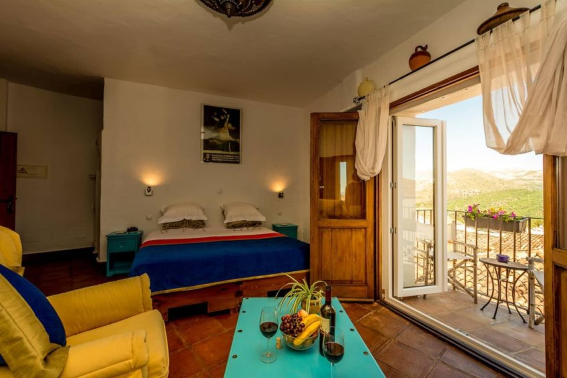 A cozy bedroom with a blue and white bed, yellow armchair, turquoise table, and balcony view of hills.