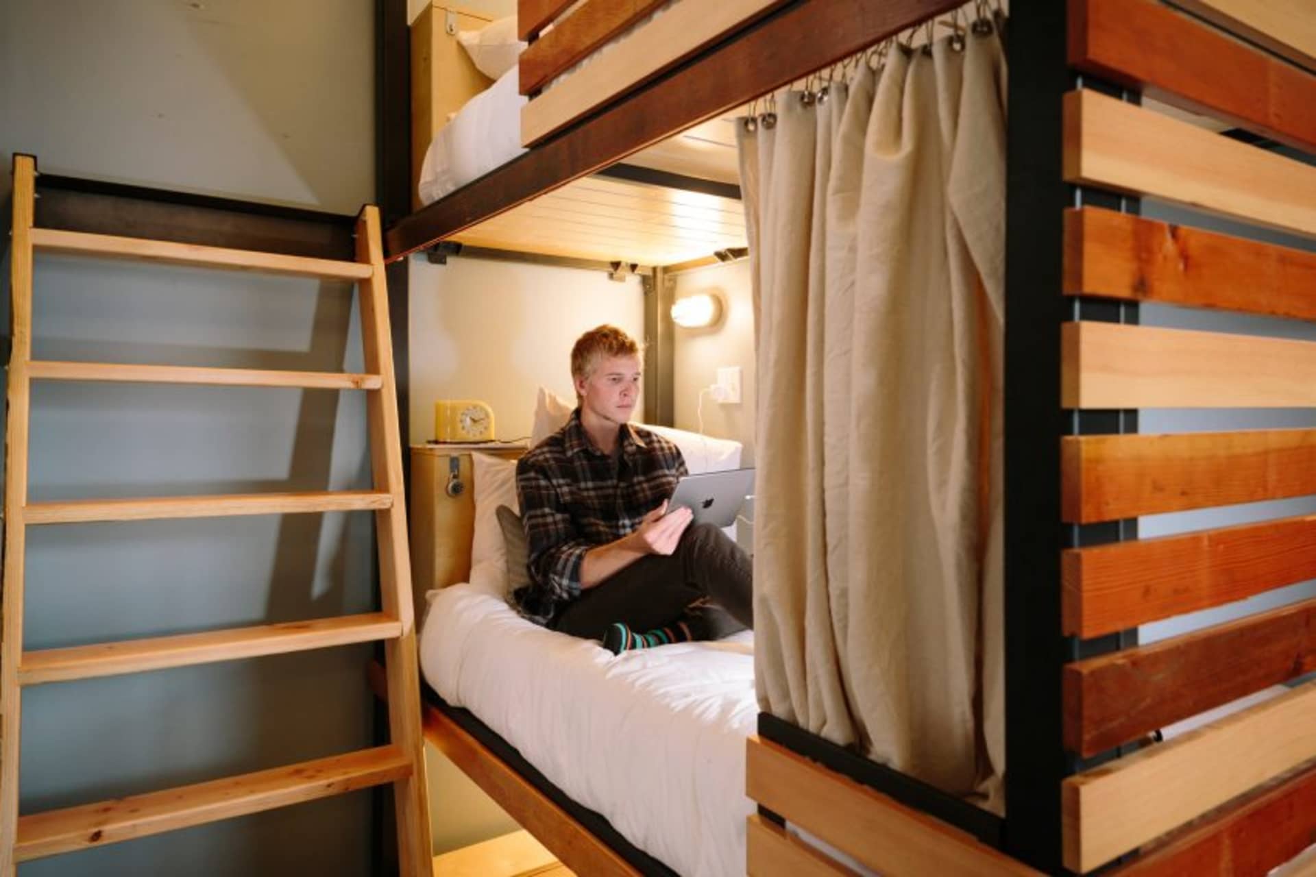 A young man sitting on a bunk bed with a laptop in a modern bedroom with wooden bunk beds and a curtain.