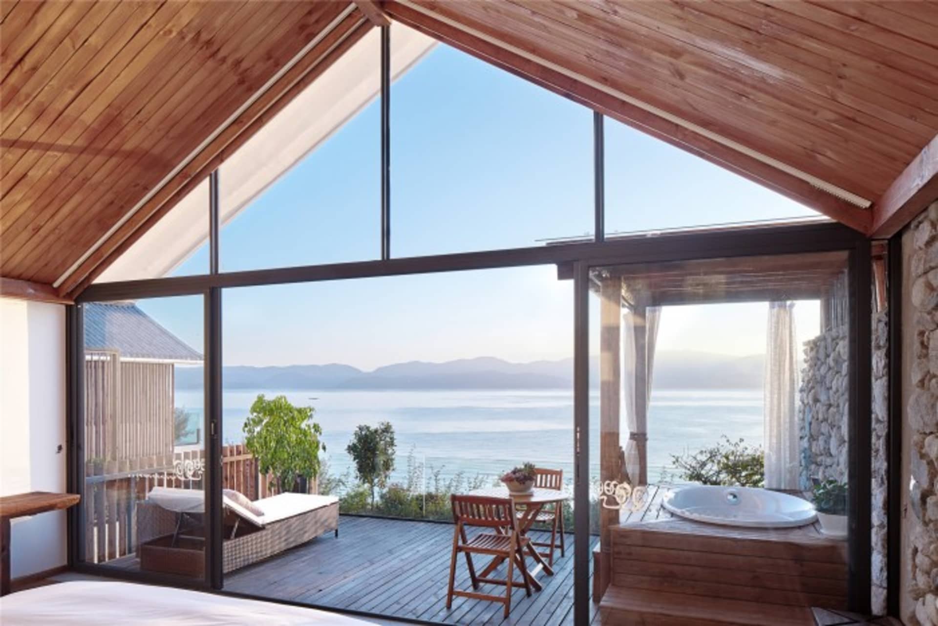 A spacious residential interior featuring a wooden deck overlooking the sea, stone wall, and glass wall and window.