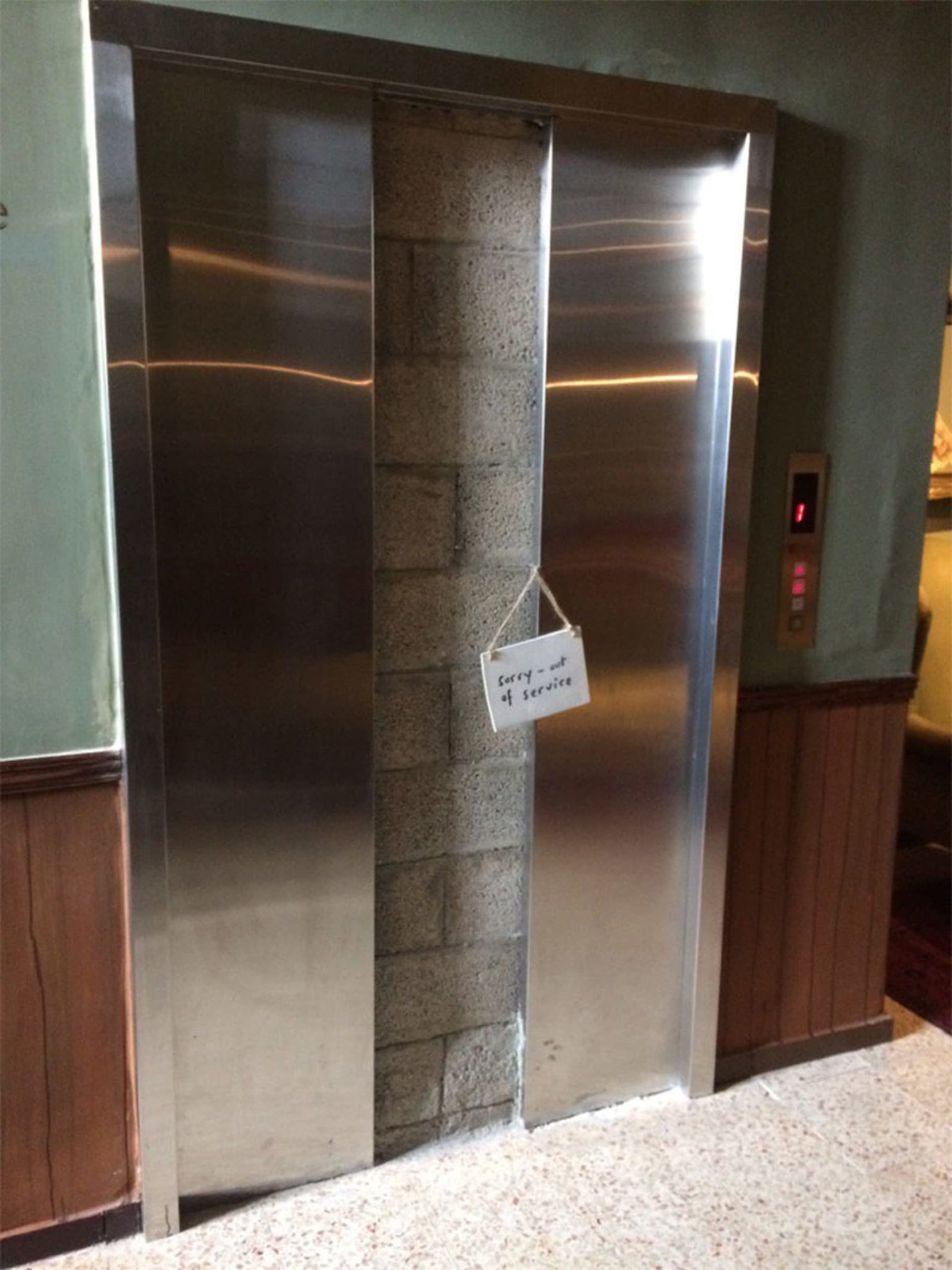 一台關閉的金屬電梯門上掛著「抱歉－停用中」的告示牌，位於綠色牆面的商業空間內