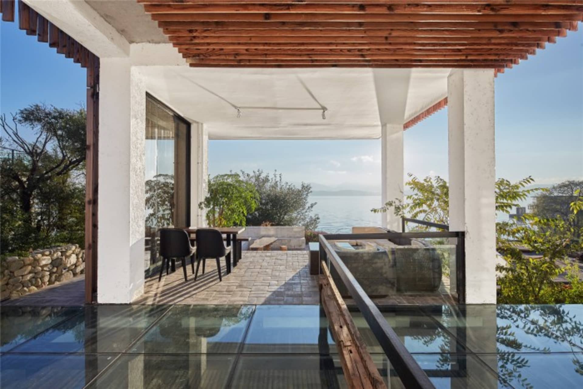 An outdoor space featuring a minimalist residential patio with wooden pergola, glass flooring, and stone elements.