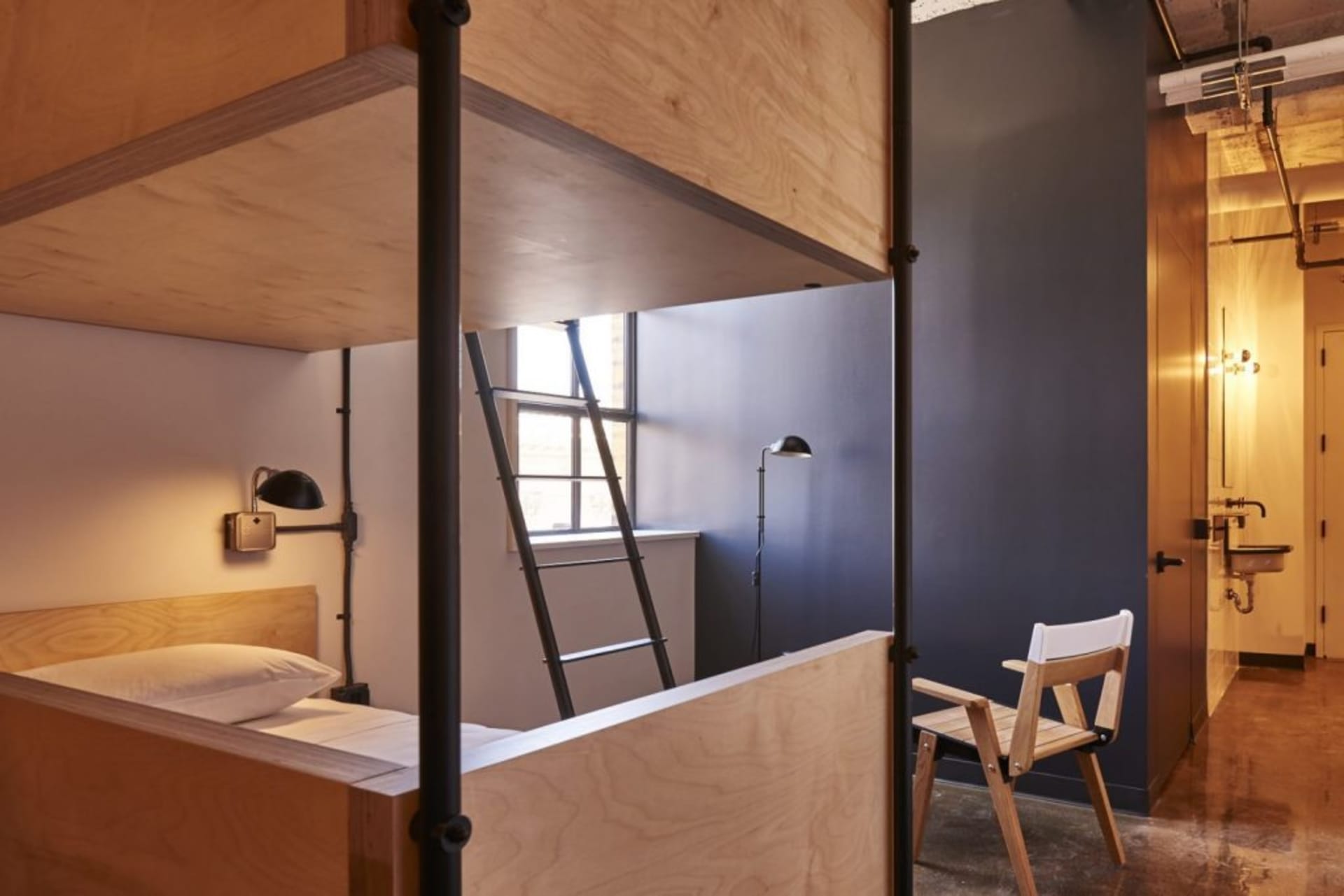 A modern bunk bed with a wooden frame and ladder, featuring a bed with a pillow on the lower bunk and a chair in the foregrou