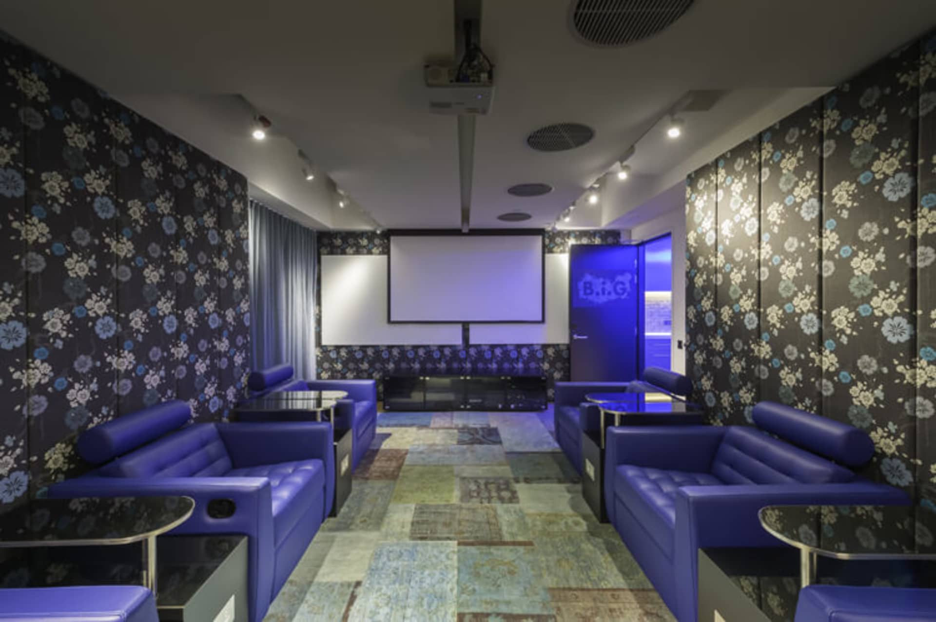 Modern recreation room featuring blue sofas on a patterned floor with a large screen and blue lighting.
