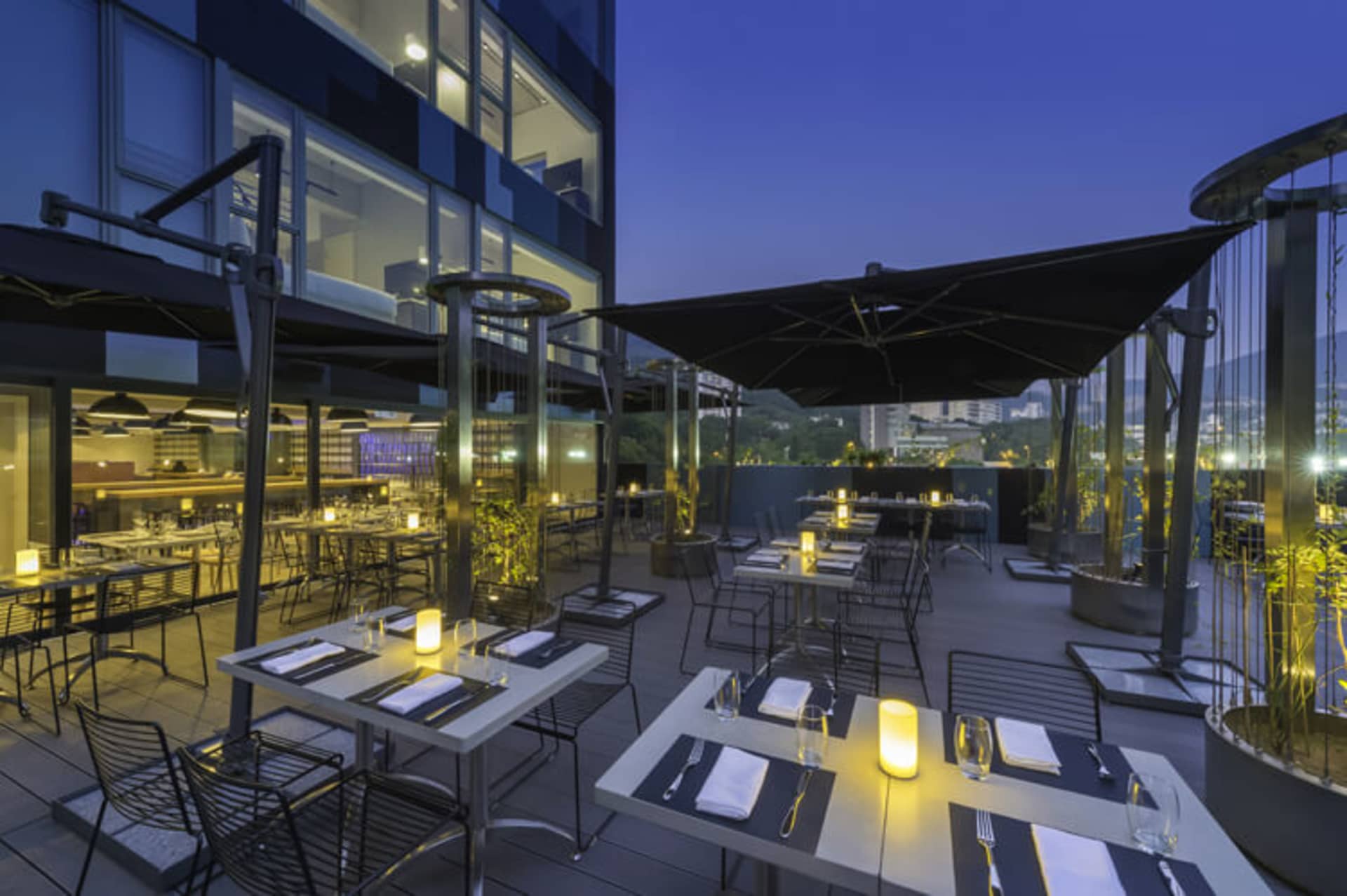 Outdoor seating area with metal furniture, glass railings, and a concrete floor, featuring tables set with dinnerware and can