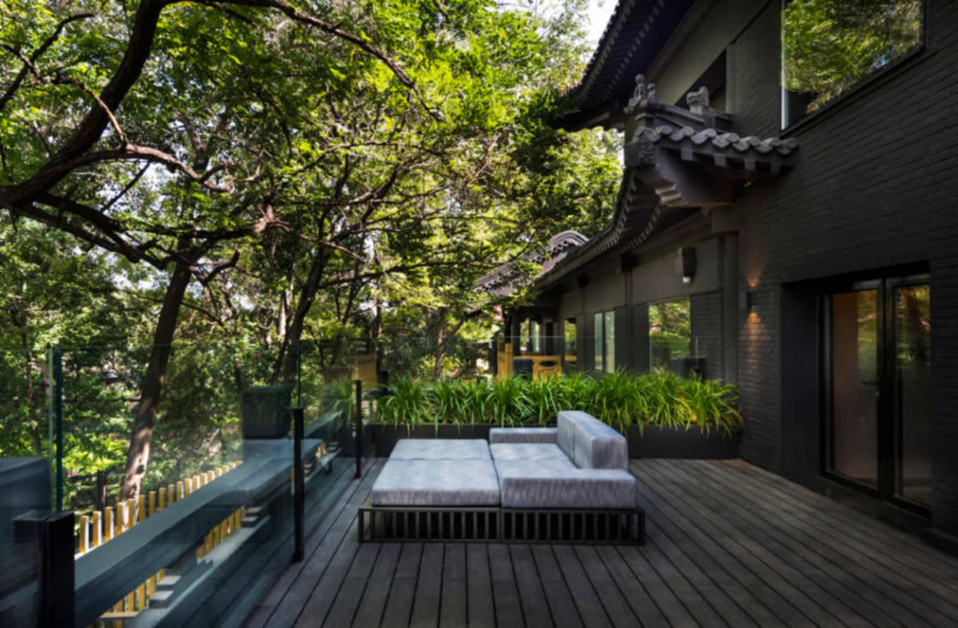 Modern residential exterior featuring wood decking, glass walls, and lush greenery.