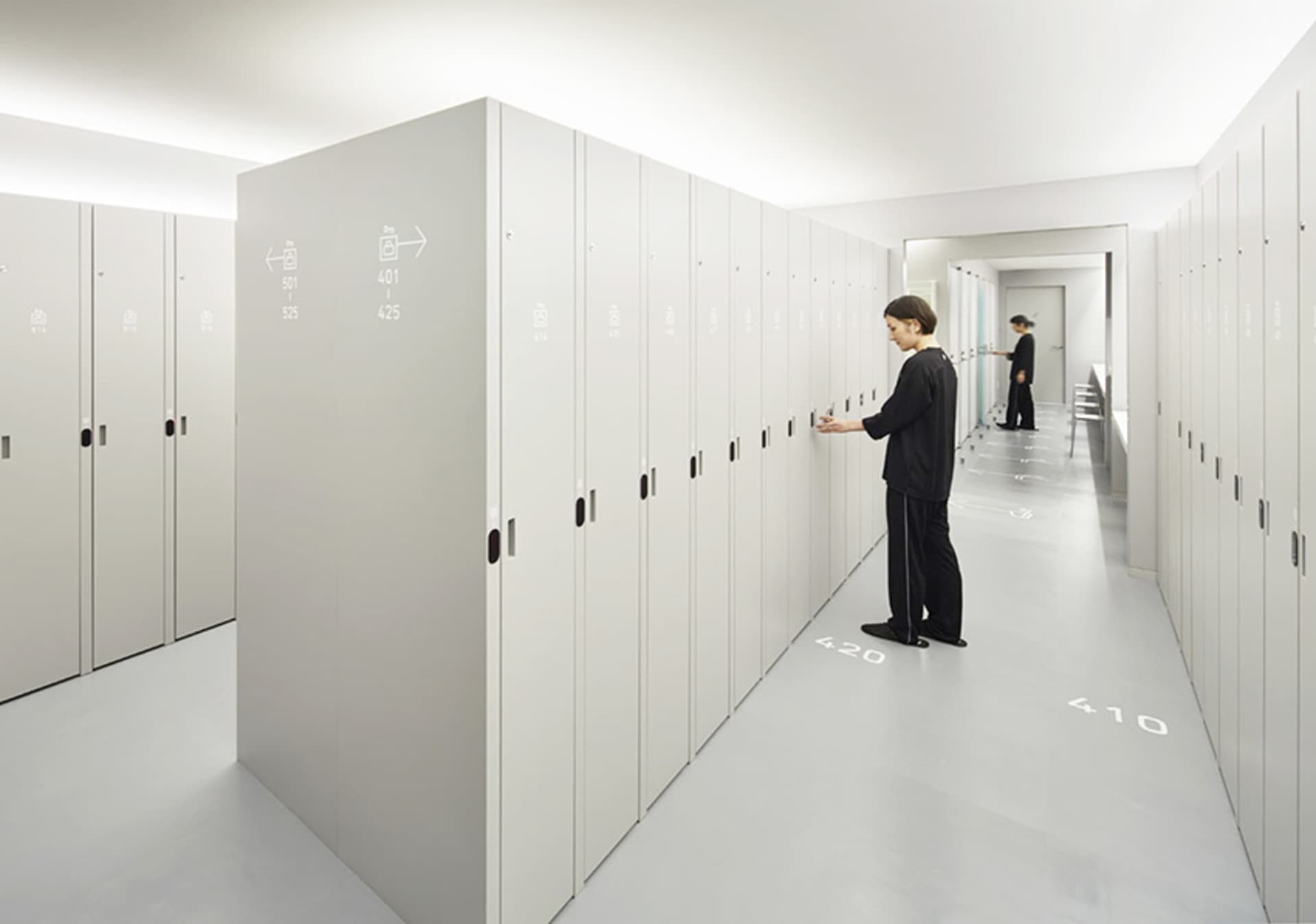 A minimalist office space featuring metal lockers with numbered labels on a white floor and walls.