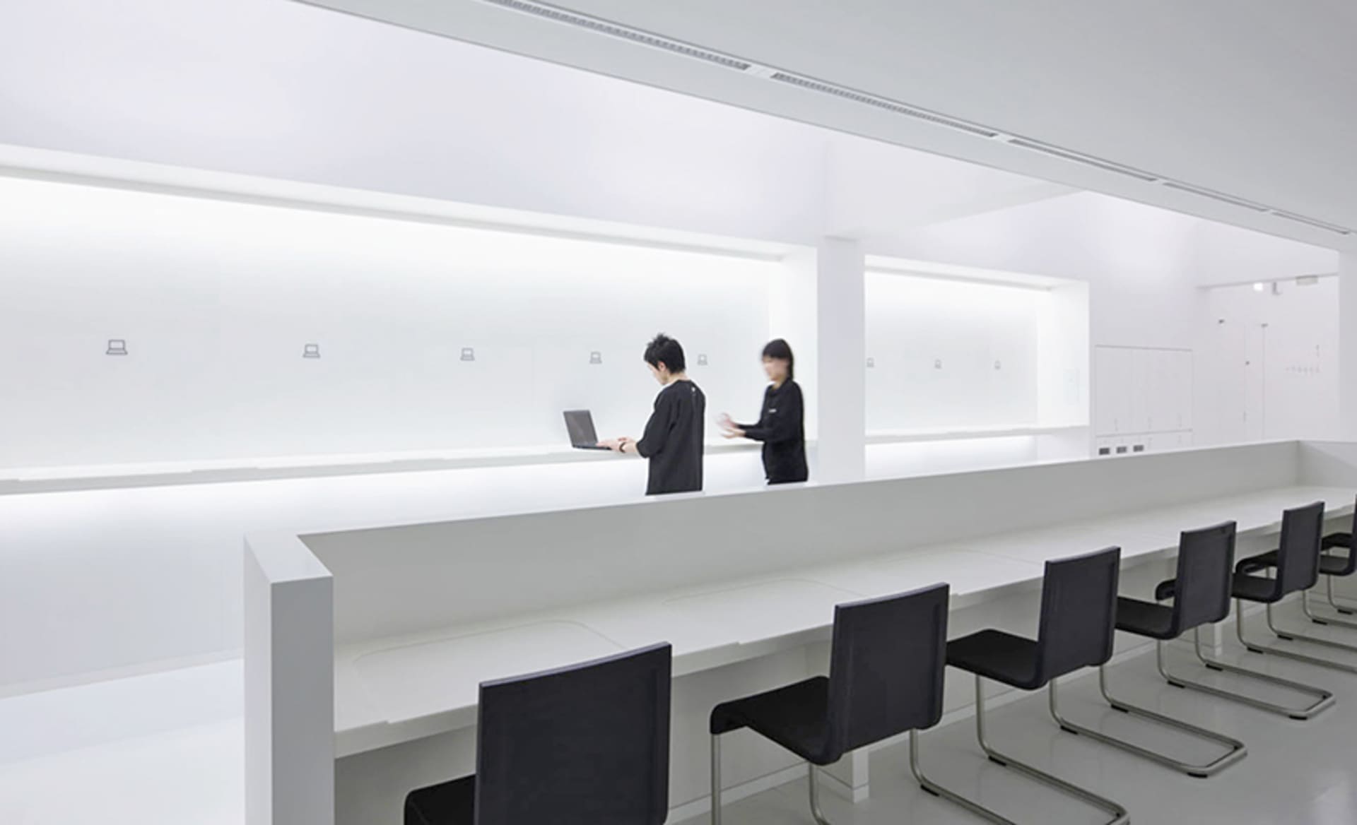 A minimalist office space with white walls, featuring open seating areas and a person working on a laptop.