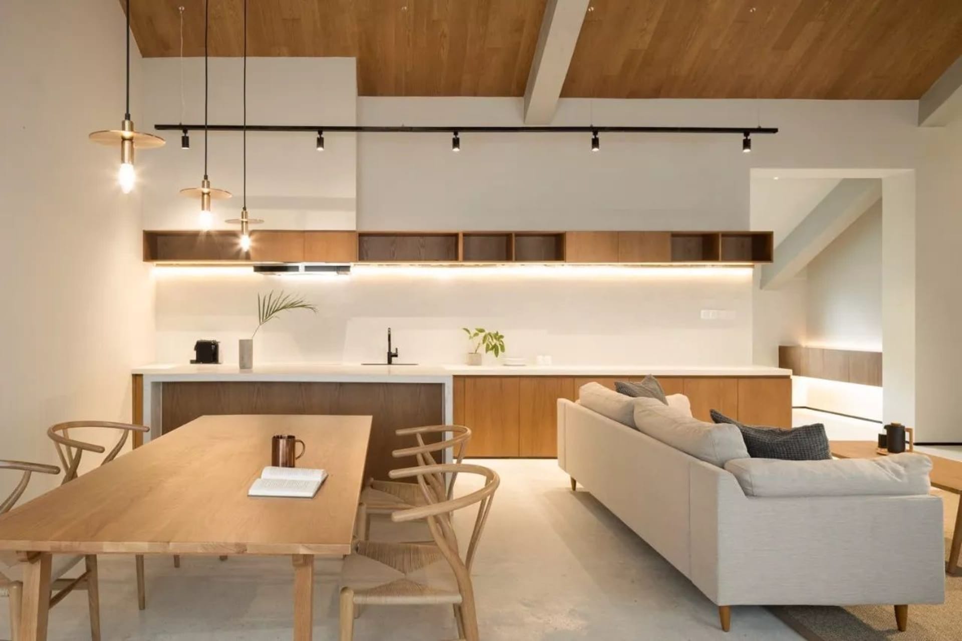 A minimalist living room featuring a wood dining table and chairs, a white sofa, and a modern kitchen area with wooden elemen