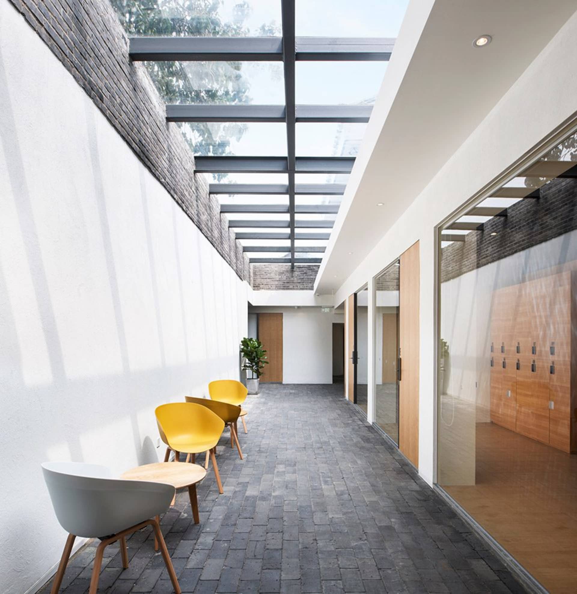 A modern corridor with glass ceiling, concrete walls, and brick flooring, featuring yellow and white chairs and a green plant