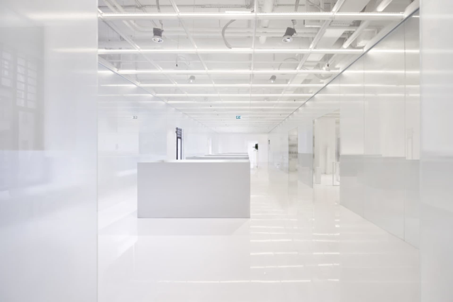 A long, white, minimalist office hallway with glass walls and overhead lighting.