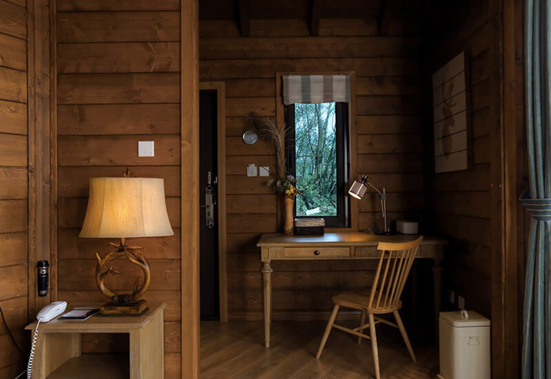 A cozy wooden home office desk with a lamp, chair, and natural light from a window.