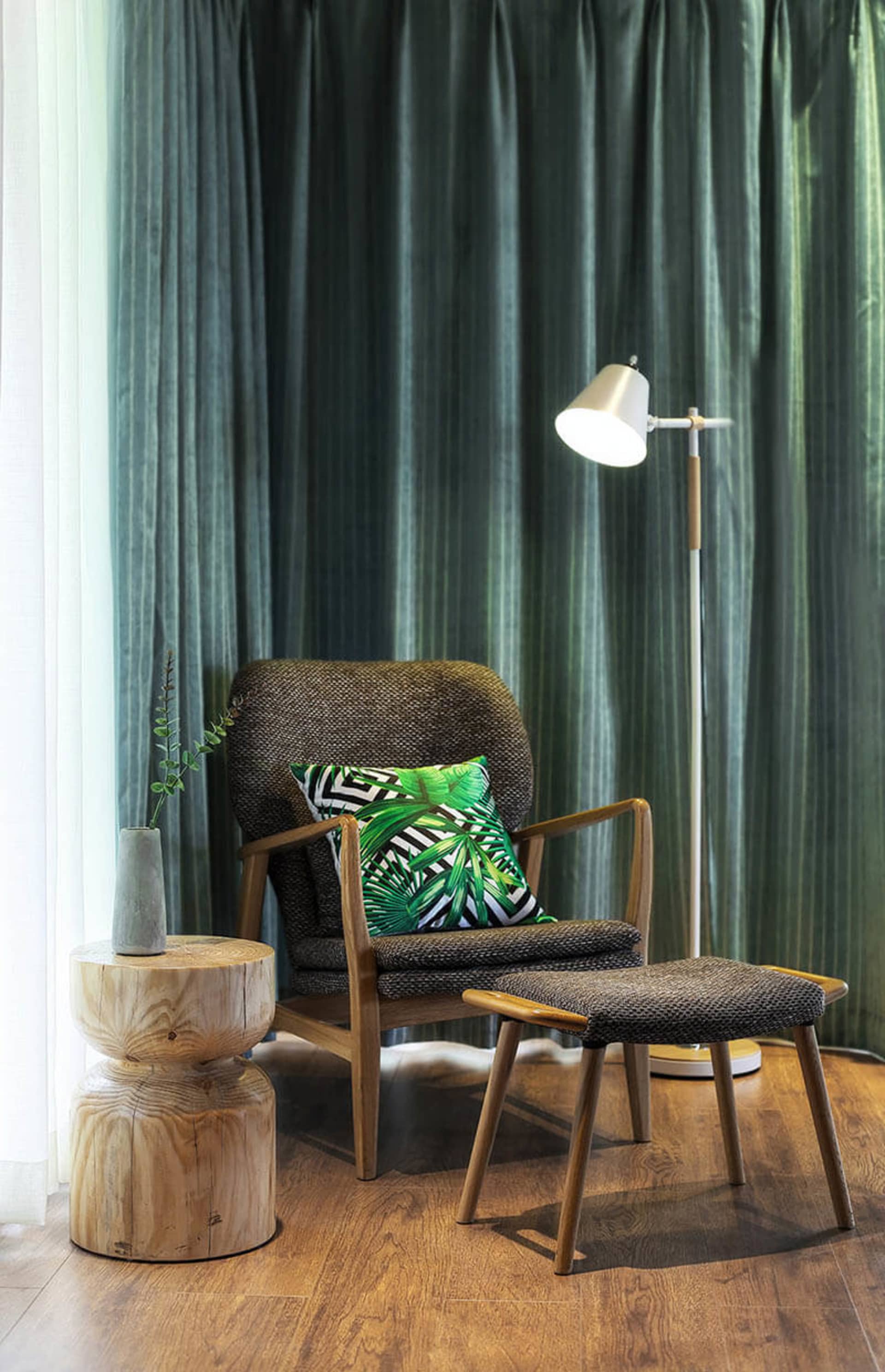 A modern living room with a comfortable chair and ottoman, featuring a palm leaf-patterned pillow and a floor lamp.