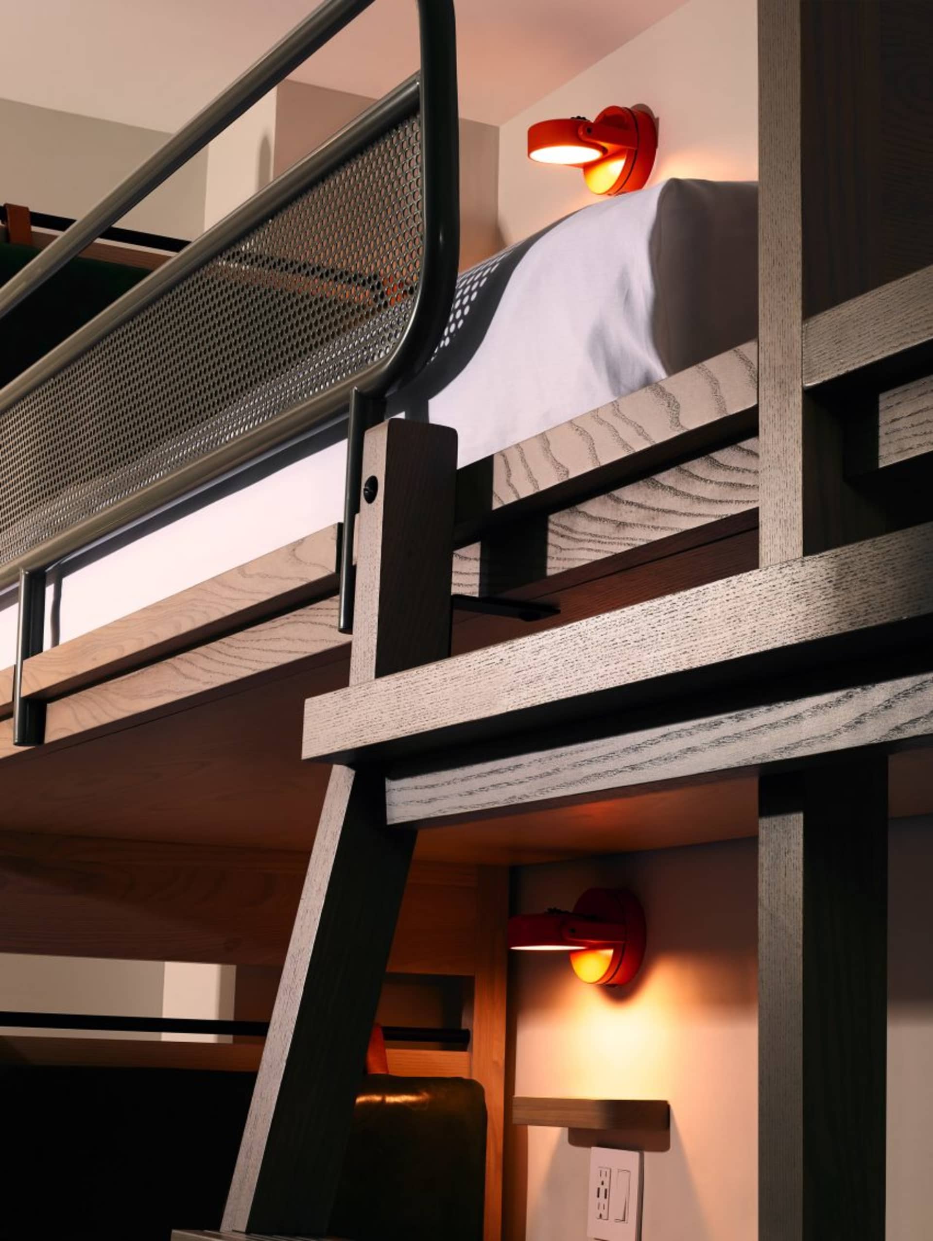 A minimalist, industrial-style bedroom featuring a wooden bunk bed with cabinets underneath and a modern red lamp.