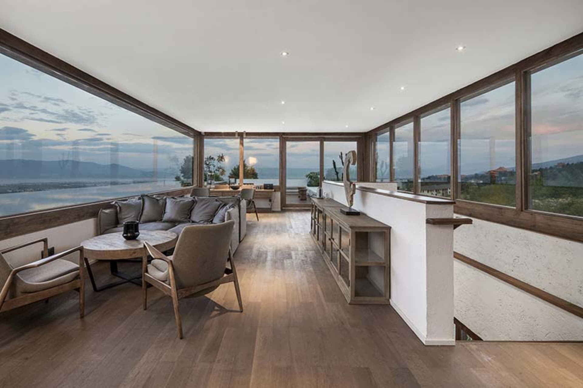 A modern living room with wooden flooring, a glass wall, and comfortable seating.