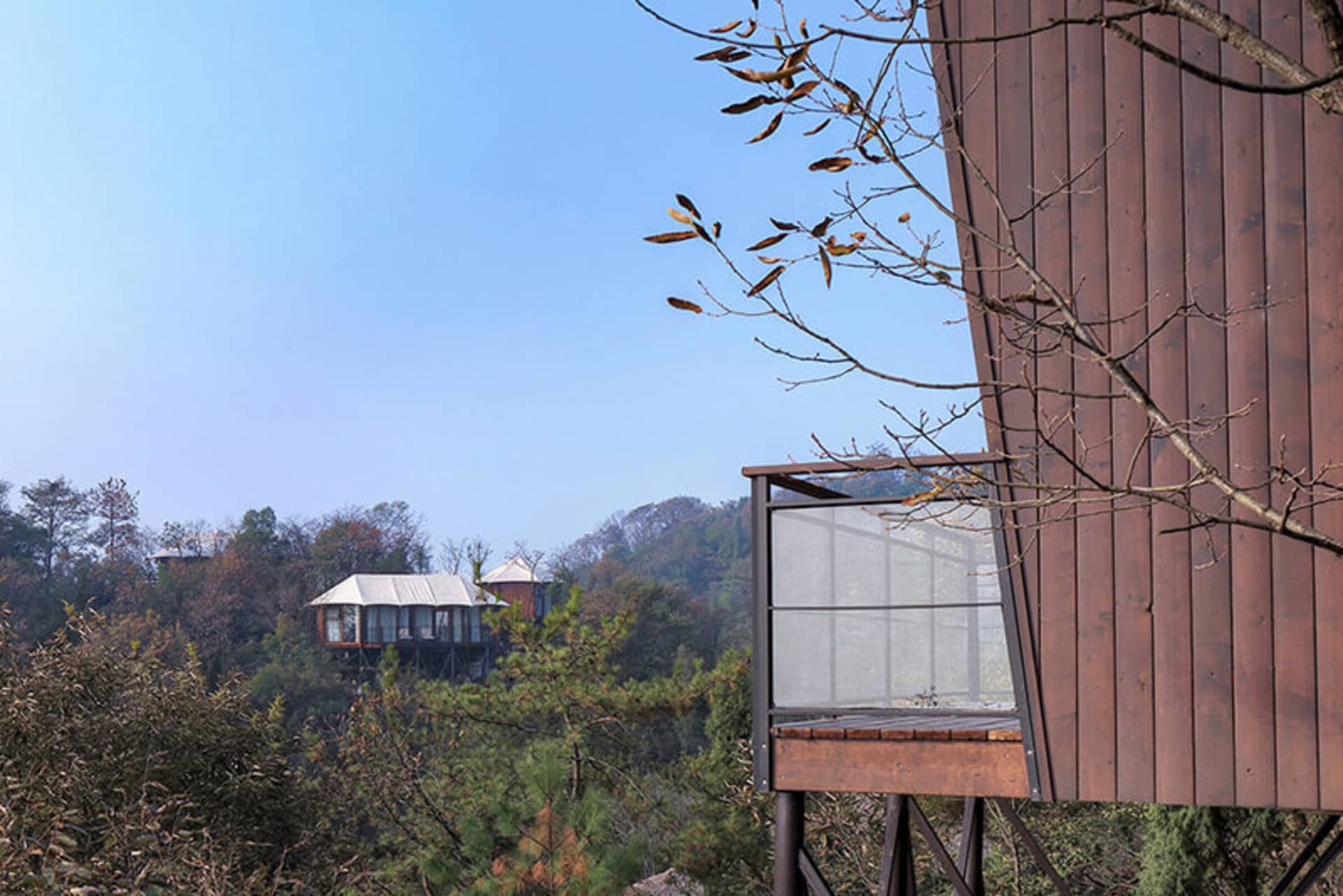 A modern, minimalist treehouse made of wood, elevated on stilts amidst lush greenery and forested mountains.