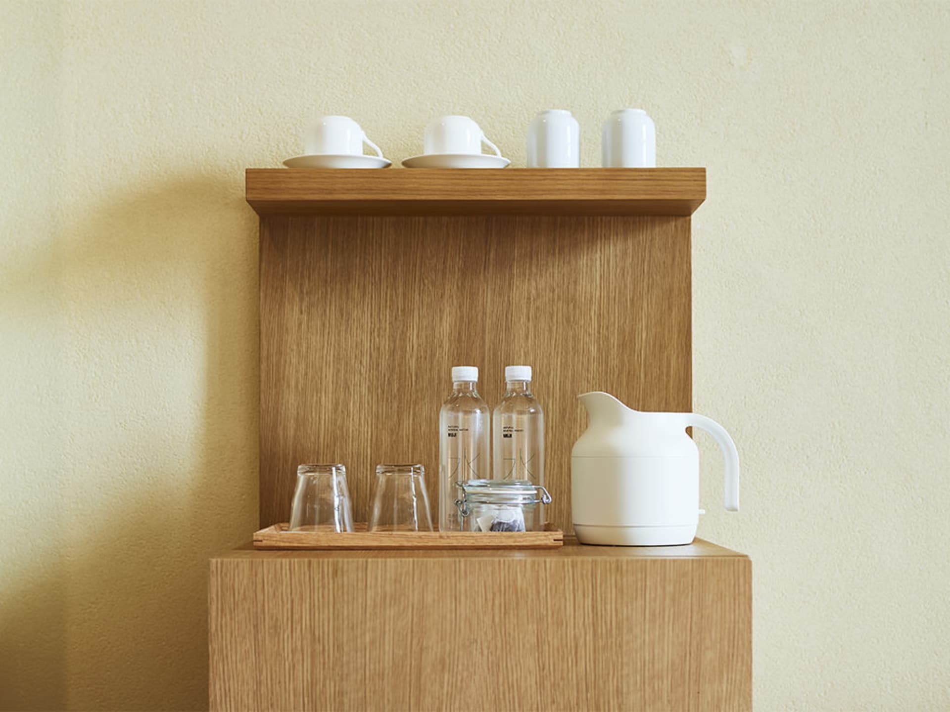 Minimalist office kitchen break room wooden shelf with white mugs, water bottles, and a white kettle.