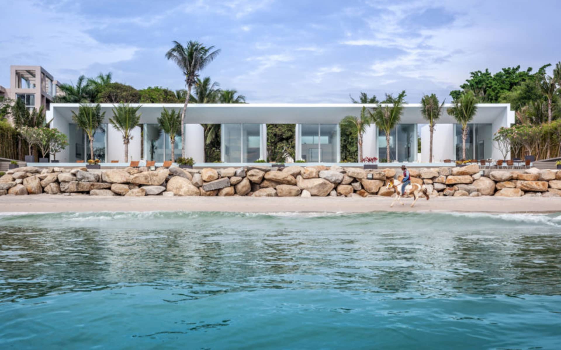 A modern beach house featuring glass walls and concrete columns, situated on a sandy shore with palm trees and blue ocean wat