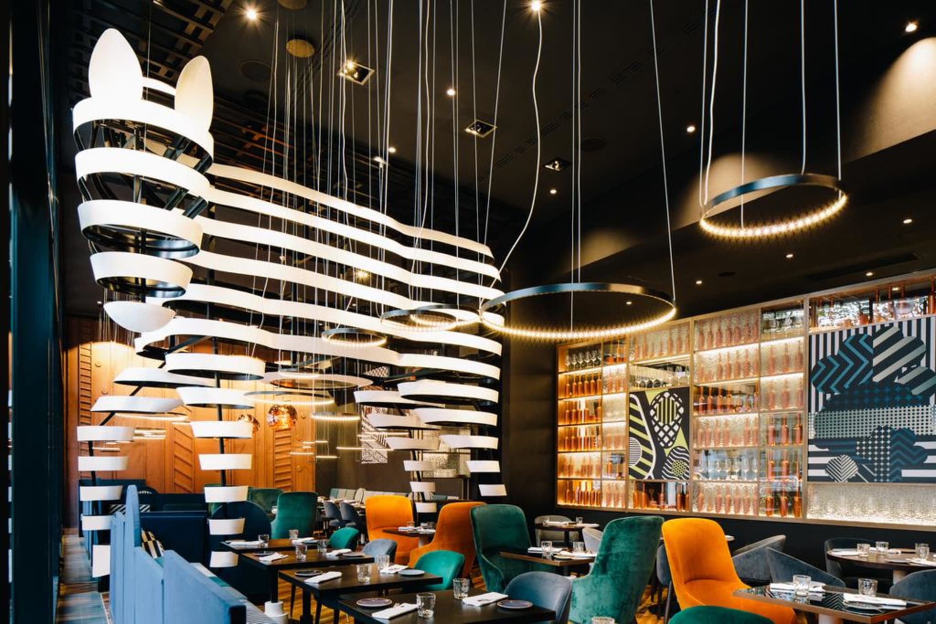 A modern restaurant interior with hanging lights, colorful chairs, and glass-fronted shelves.