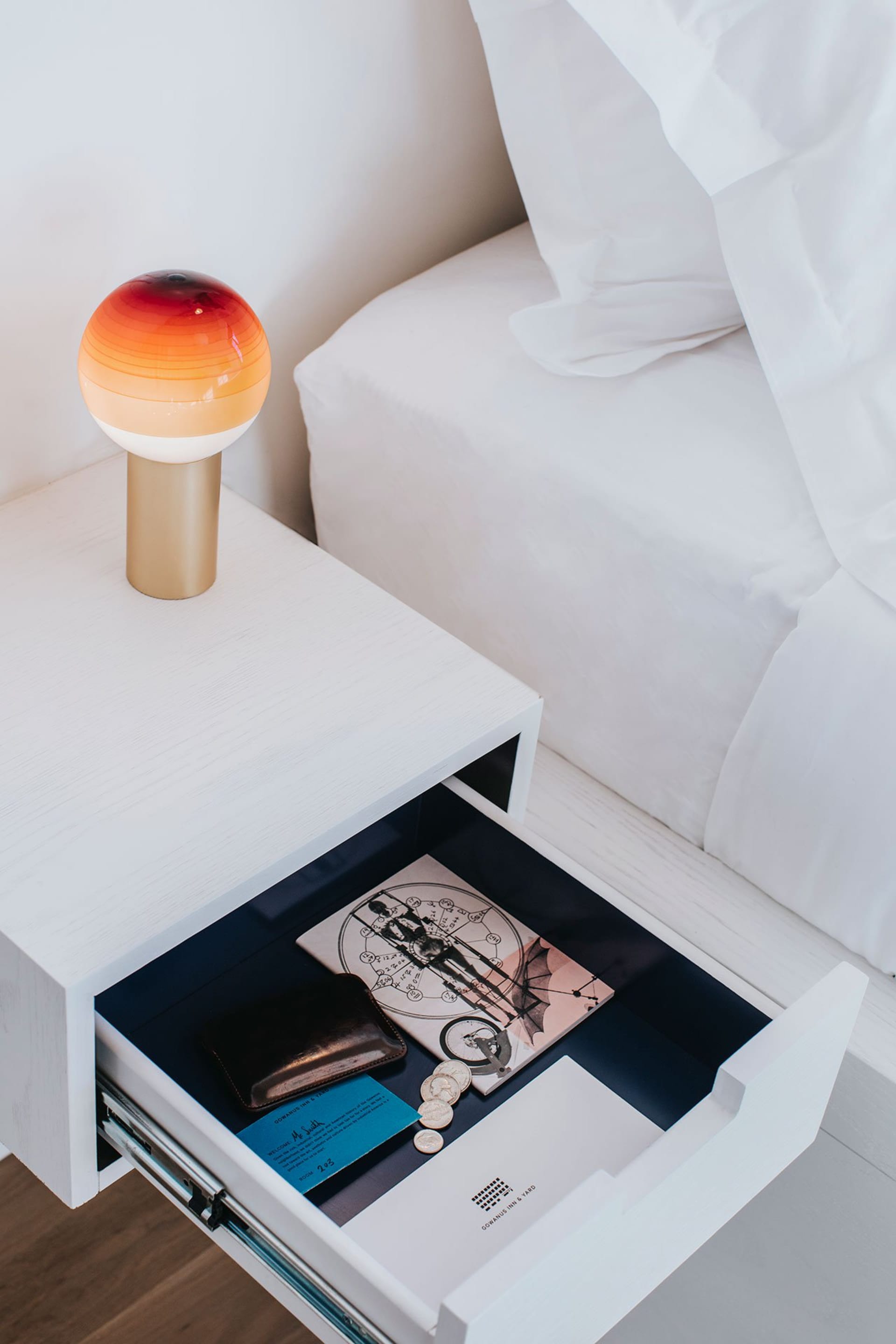 A modern white bedroom decorated with a gold and orange light, a white bed, and an open drawer containing a leather wallet, a