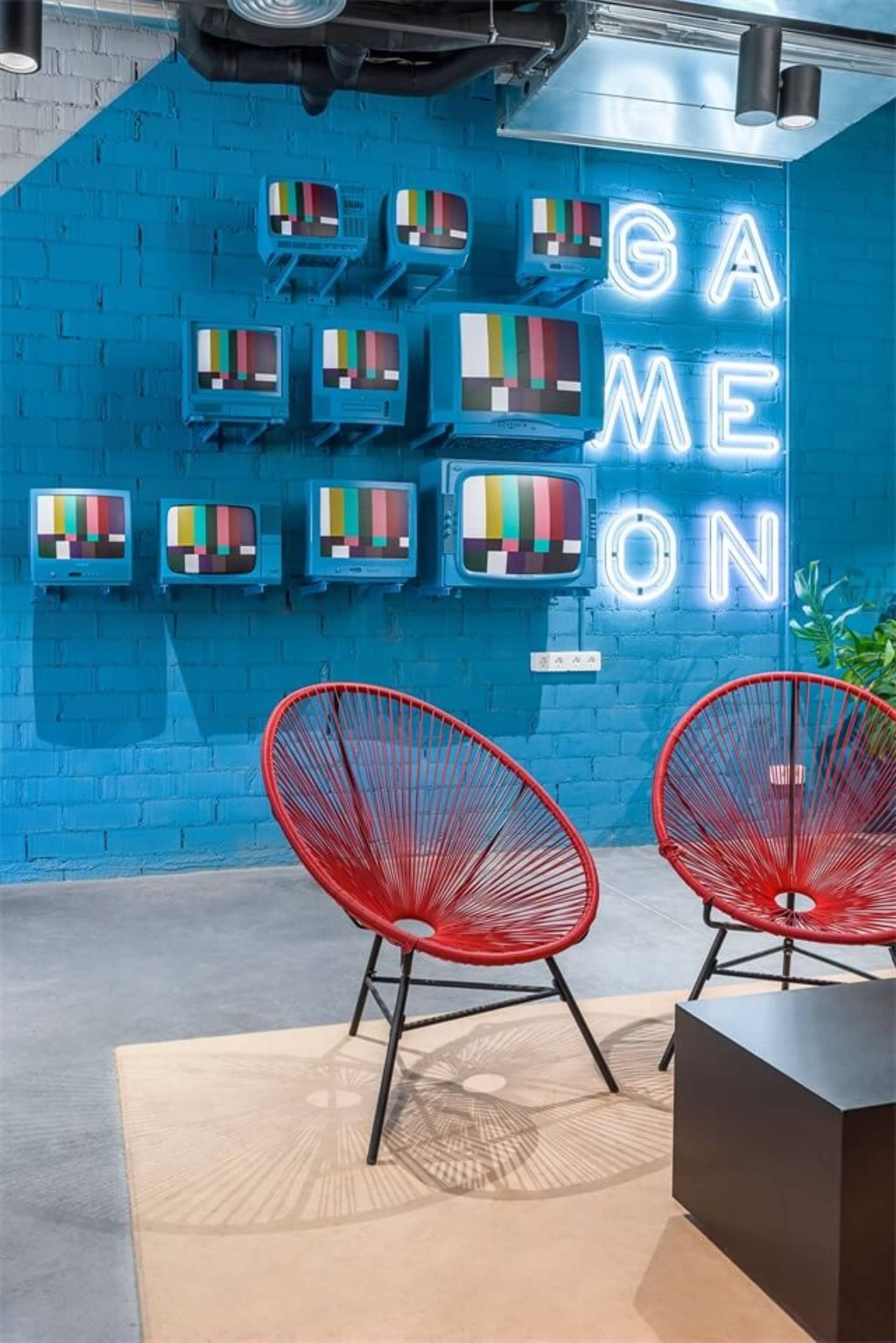 A modern lobby features red metal and fabric lounge chairs, a blue brick wall with neon sign reading "GAME ON", and a gray fl