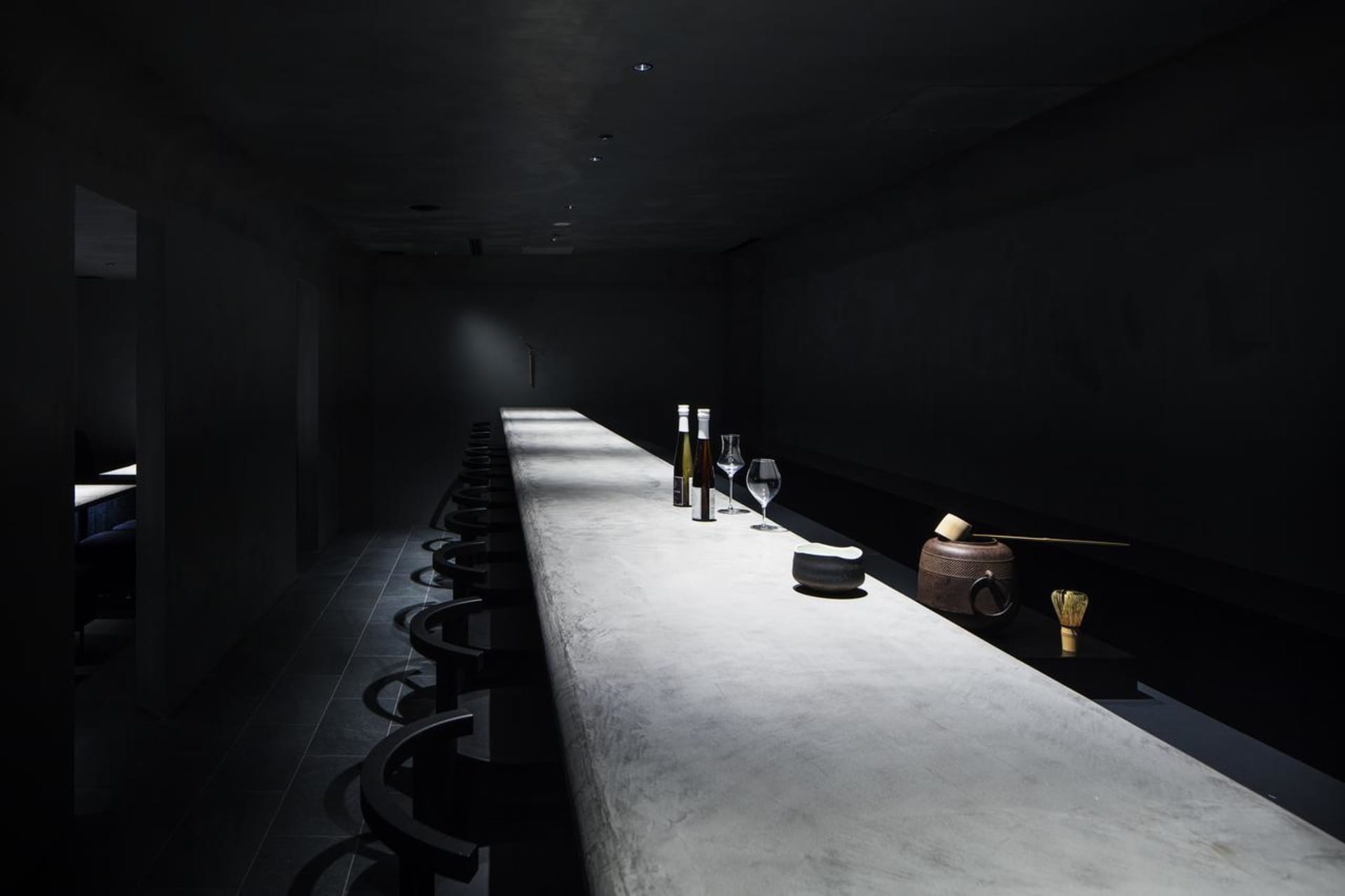A minimalist restaurant bar with a concrete countertop and black seating on a dark tiled floor.