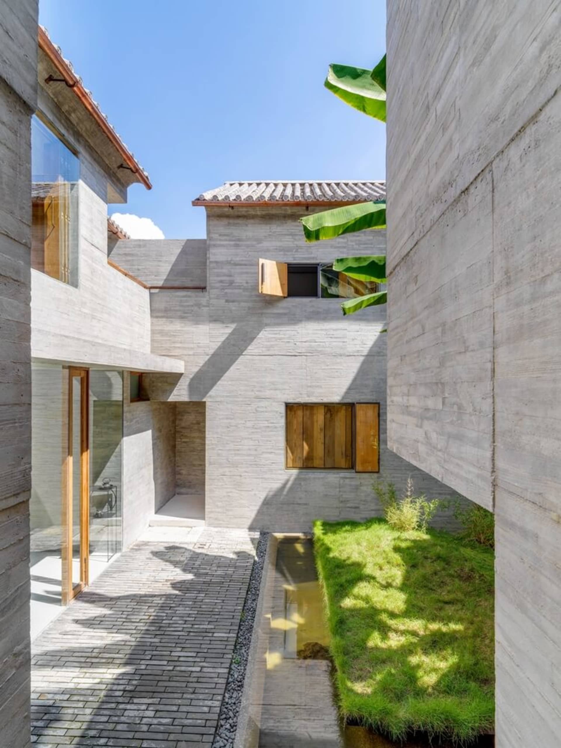 A minimalist house exterior with concrete walls, wooden window shutters, and a small garden with grass and plants, under a cl