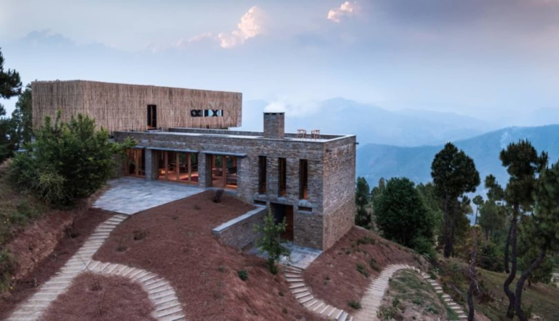 A modern house with stone and wood exterior, located on a hill with a view of the mountains and forest.