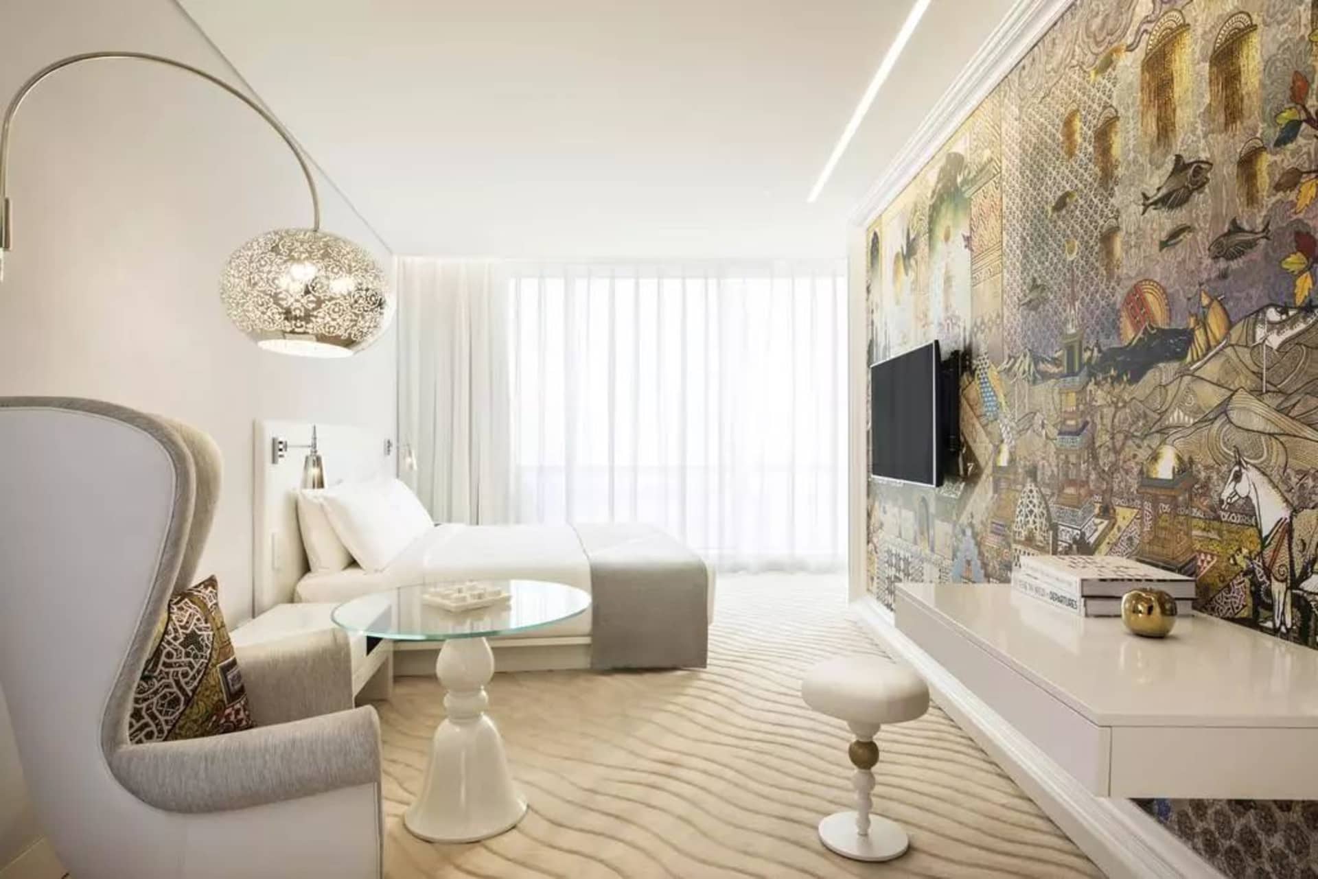 Modern bedroom featuring a bed with white linens, a white armchair with decorative accents, a glass coffee table, and a decor