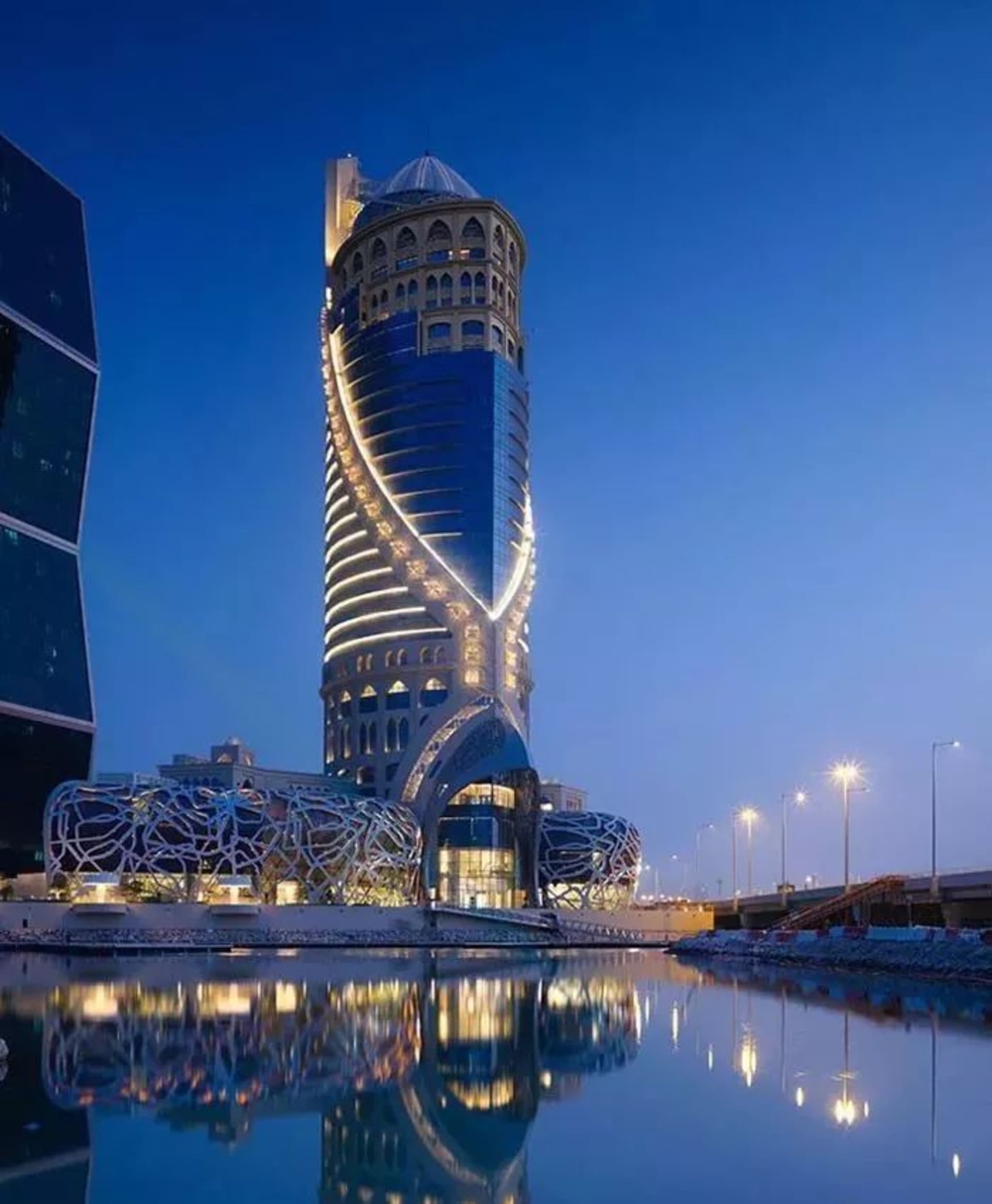 A modern, spiral-shaped building made of glass, concrete, and metal materials, reflecting in a nearby body of water.