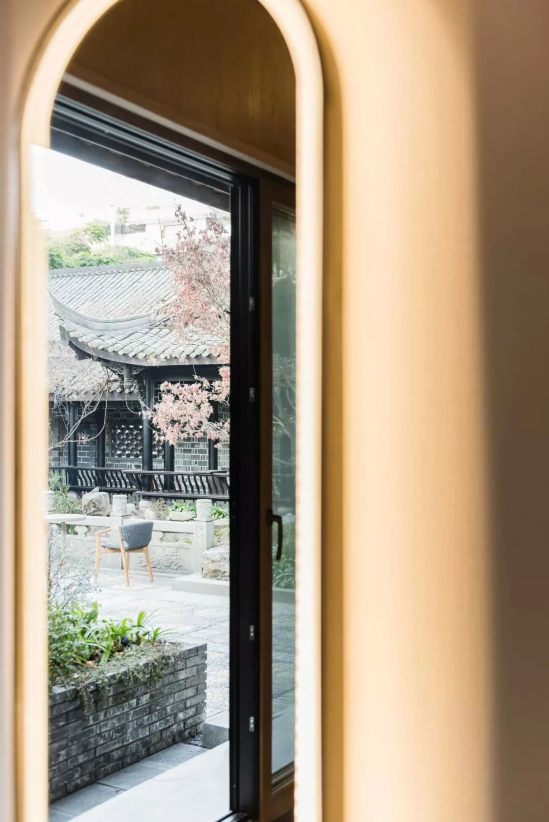 透過拱形窗戶眺望庭院，庭院內有椅子、植物，以及一棵開滿粉色花朵的樹。