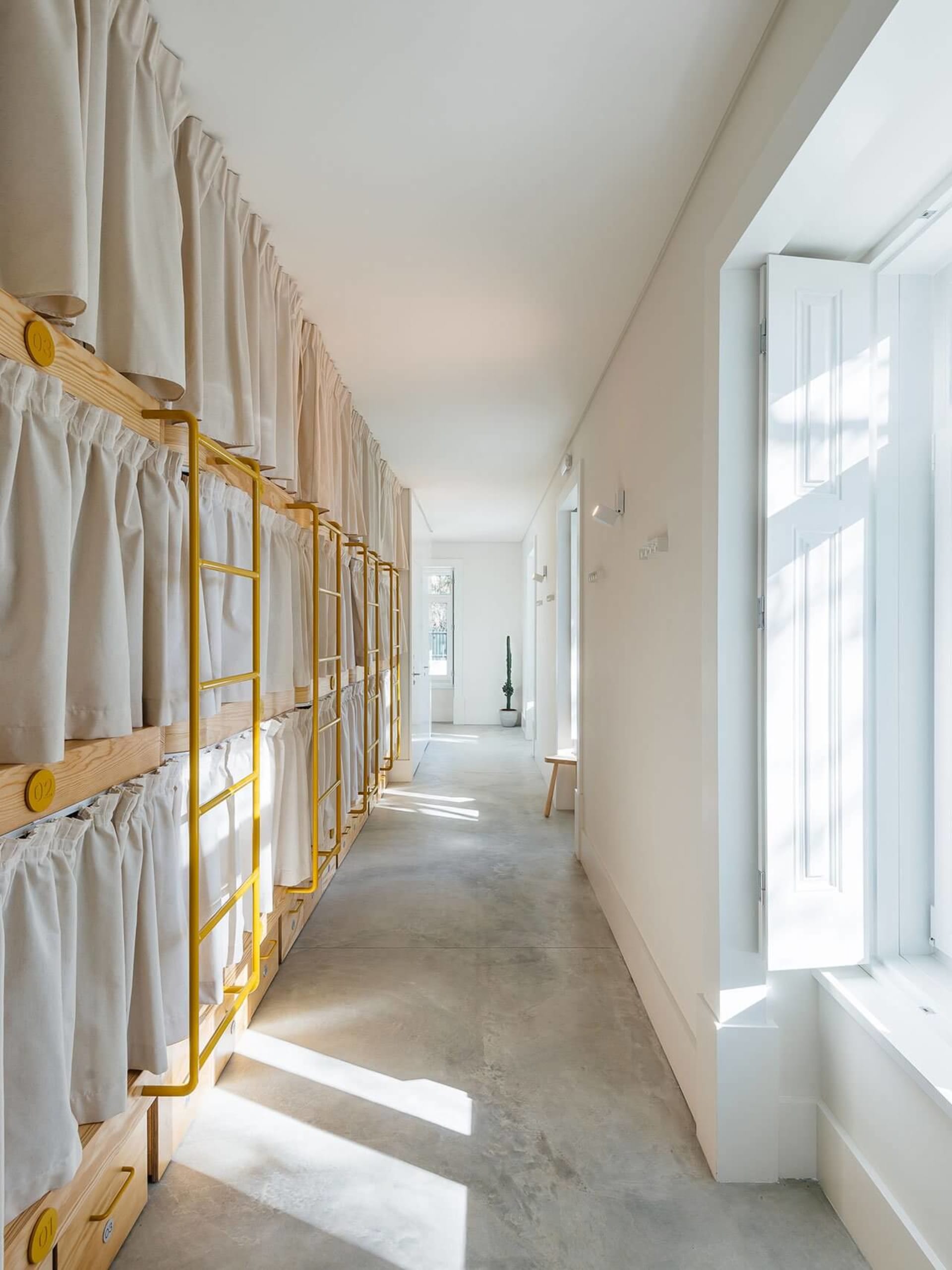 Minimalist hostel room featuring white curtains on bunk beds with yellow supports and concrete flooring.