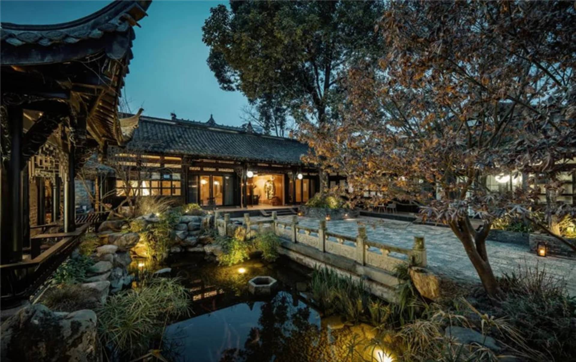 A traditional Chinese residential exterior with stone walls, wooden doors, and a stone bridge over a pond at night.