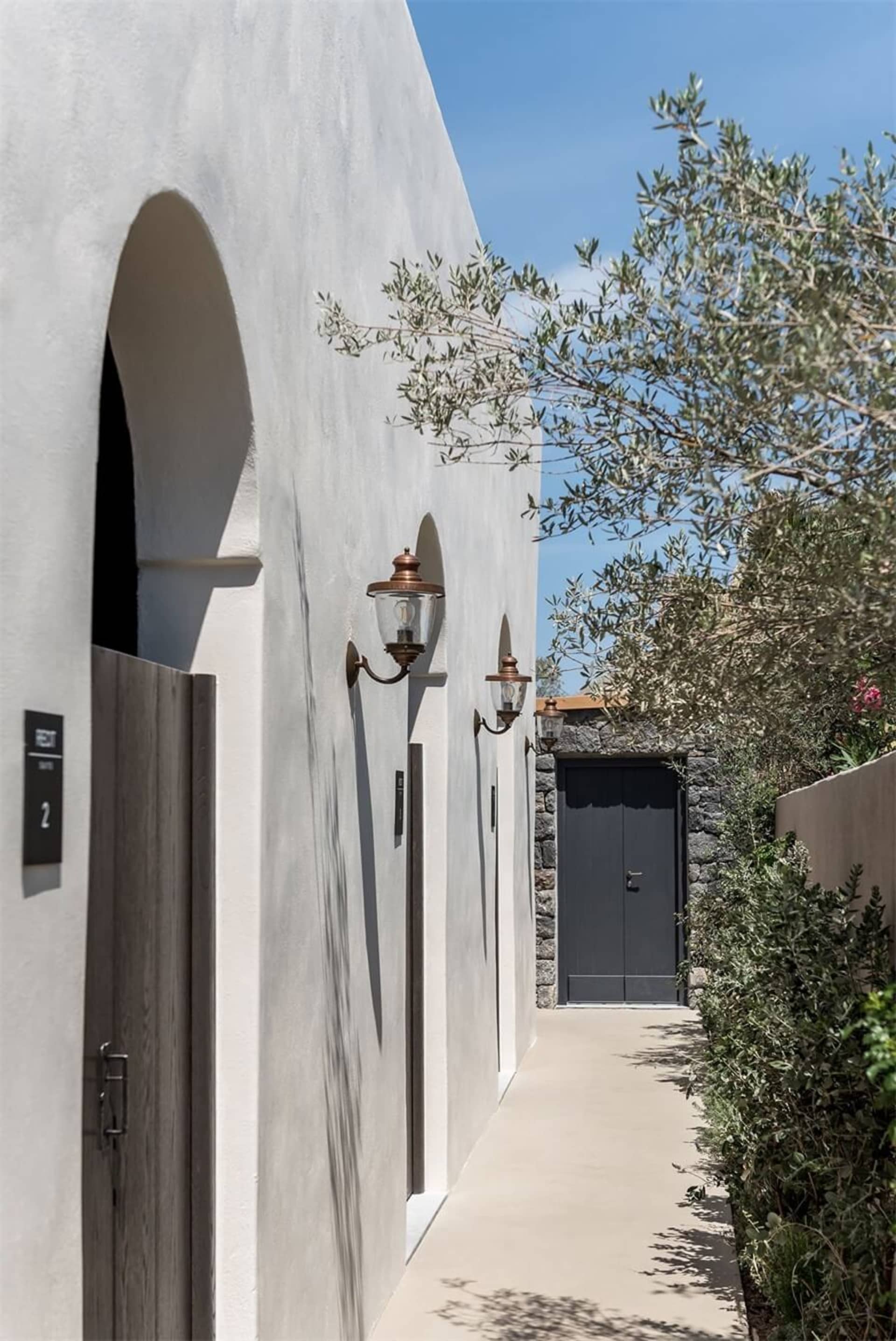 A white stucco entrance with arched doorways and wooden doors, surrounded by greenery and a clear blue sky.
