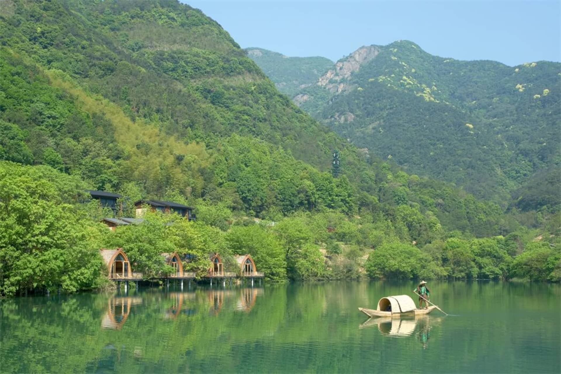 寧靜的湖畔度假村，綠意盎然，湖面停泊一艘小船，遠方群山環繞。