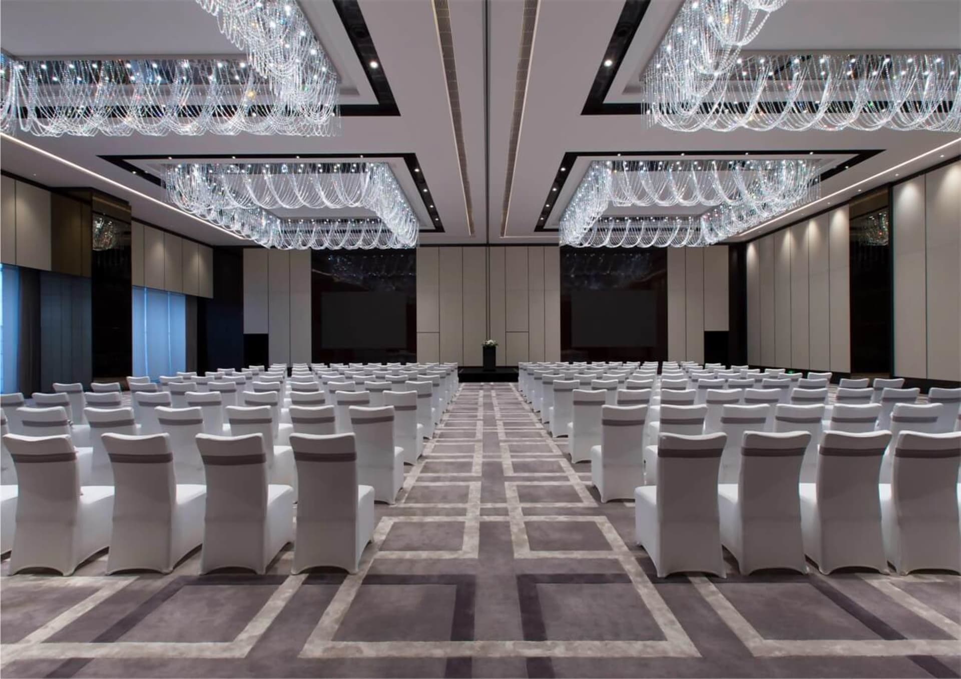 A large, well-lit conference room with white chairs covered in white fabric, carpeted with a checkered pattern, and adorned w