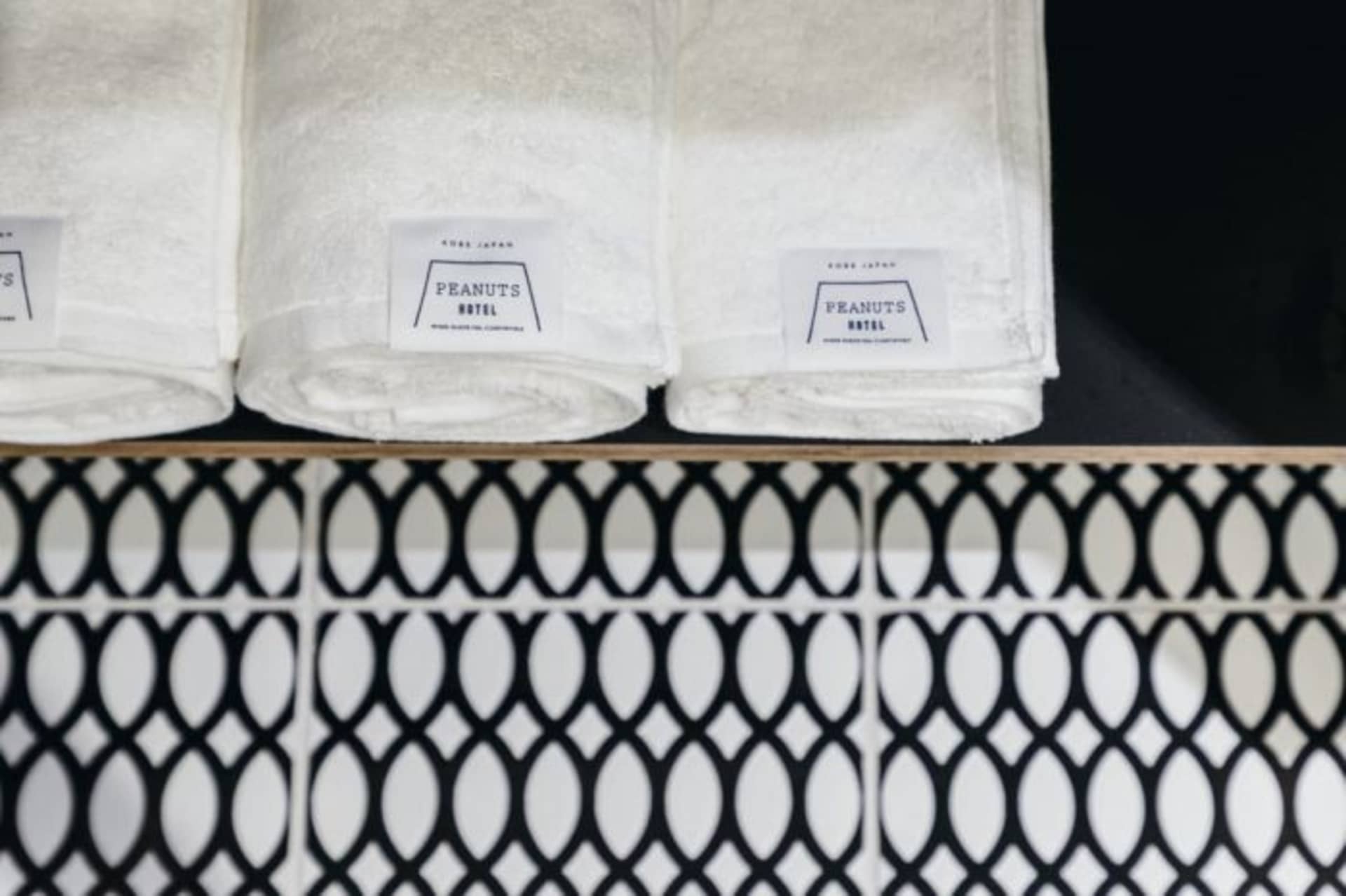 Three white towels with 'PEANUTS HOTEL' labels rolled up and placed on a shelf above a wall with black and white patterned ti