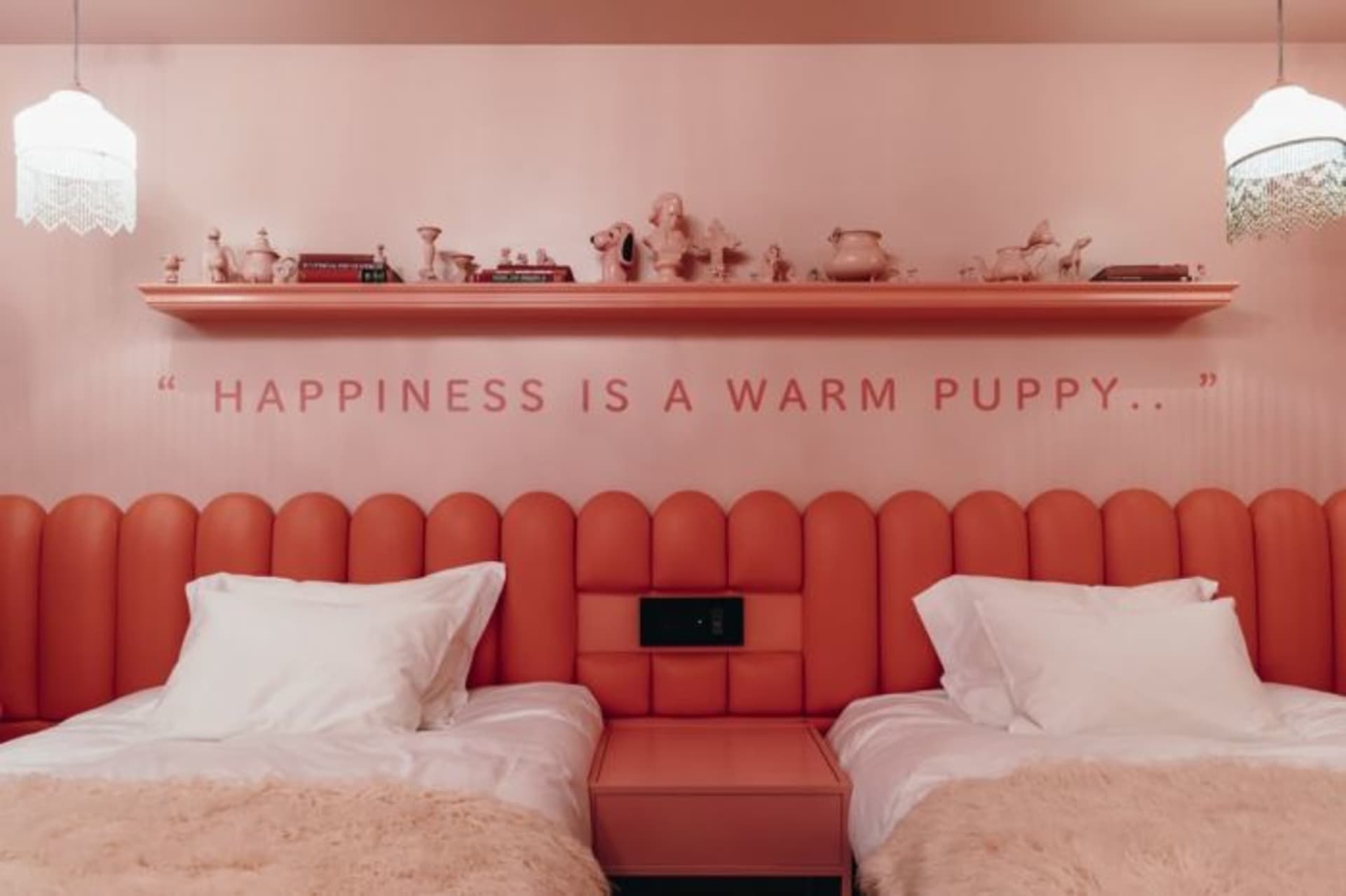 A pink and orange retro-designed bedroom with twin beds, a cushioned headboard, and a shelf displaying ceramic items.