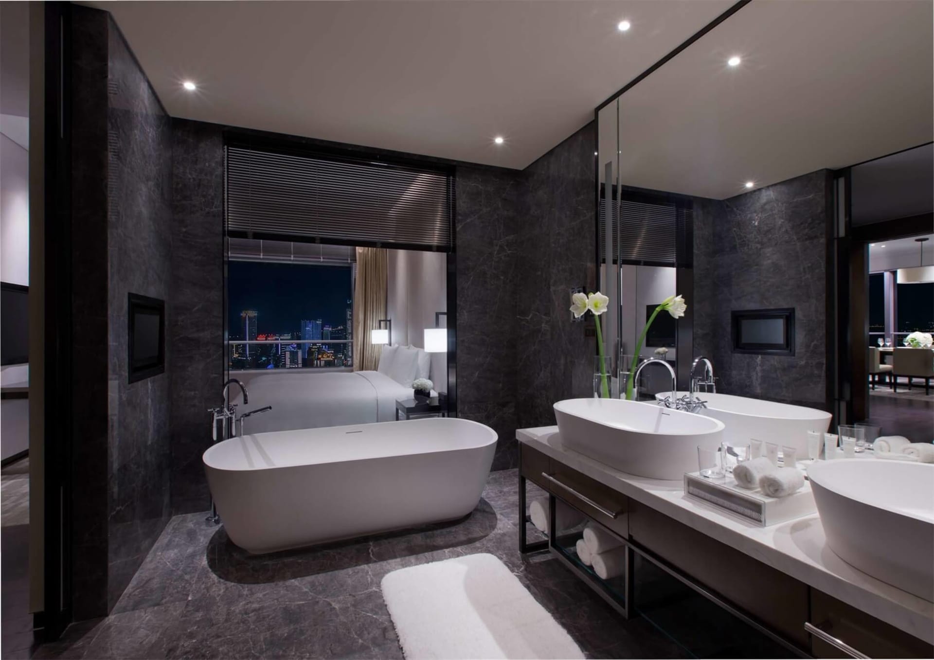 A modern bathroom with marble countertops, black granite walls, and a freestanding bathtub overlooking a cityscape.