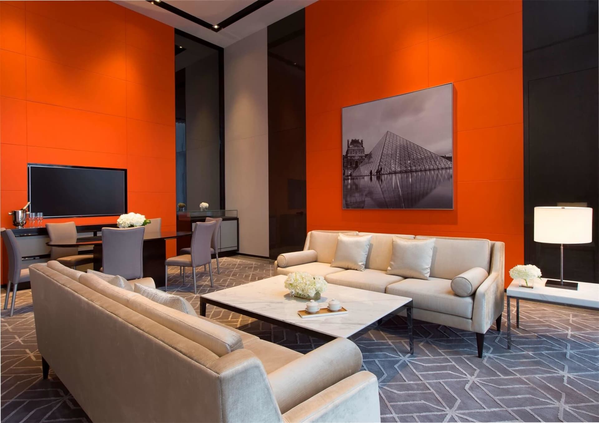 Modern minimalist lounge area with white sofas, marble coffee table, and orange accent wall featuring a black and white photo