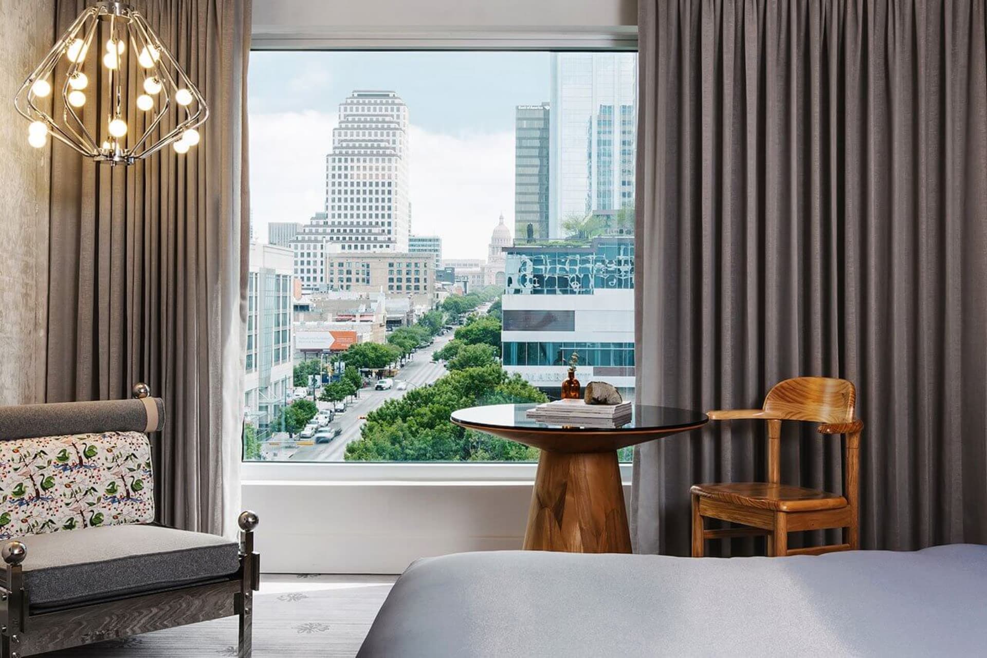 Modern minimalist hotel room with a view of the city, featuring a gray bed, a wooden chair, a round glass table, and fabric c
