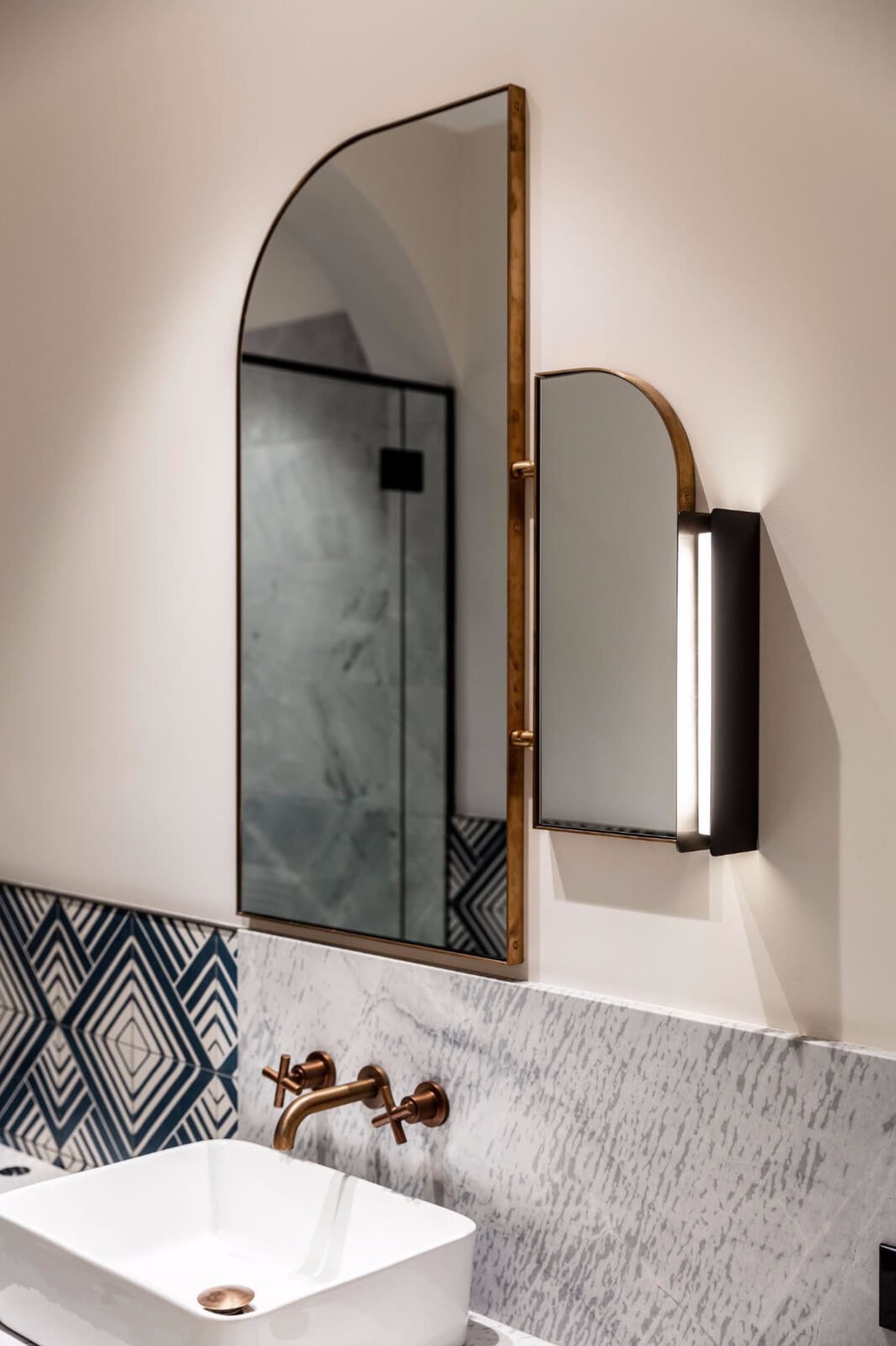 Modern bathroom featuring metallic brass accents and wall-mounted light fixture with marble tiled backdrop.
