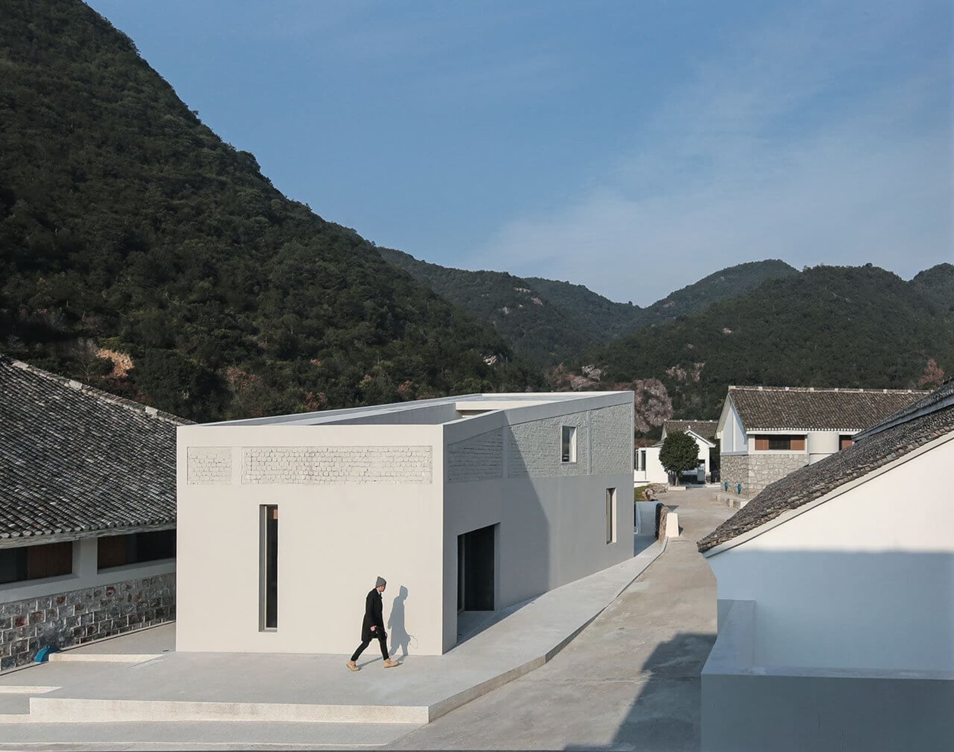 A modern building made of concrete with a person walking on a walkway in front of it, set against a backdrop of green hills a
