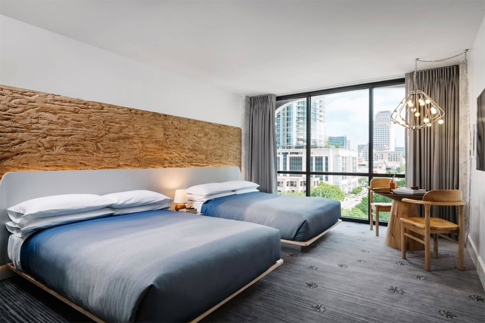 A modern hotel room with two beds, a desk, and a large window providing a view of the city.